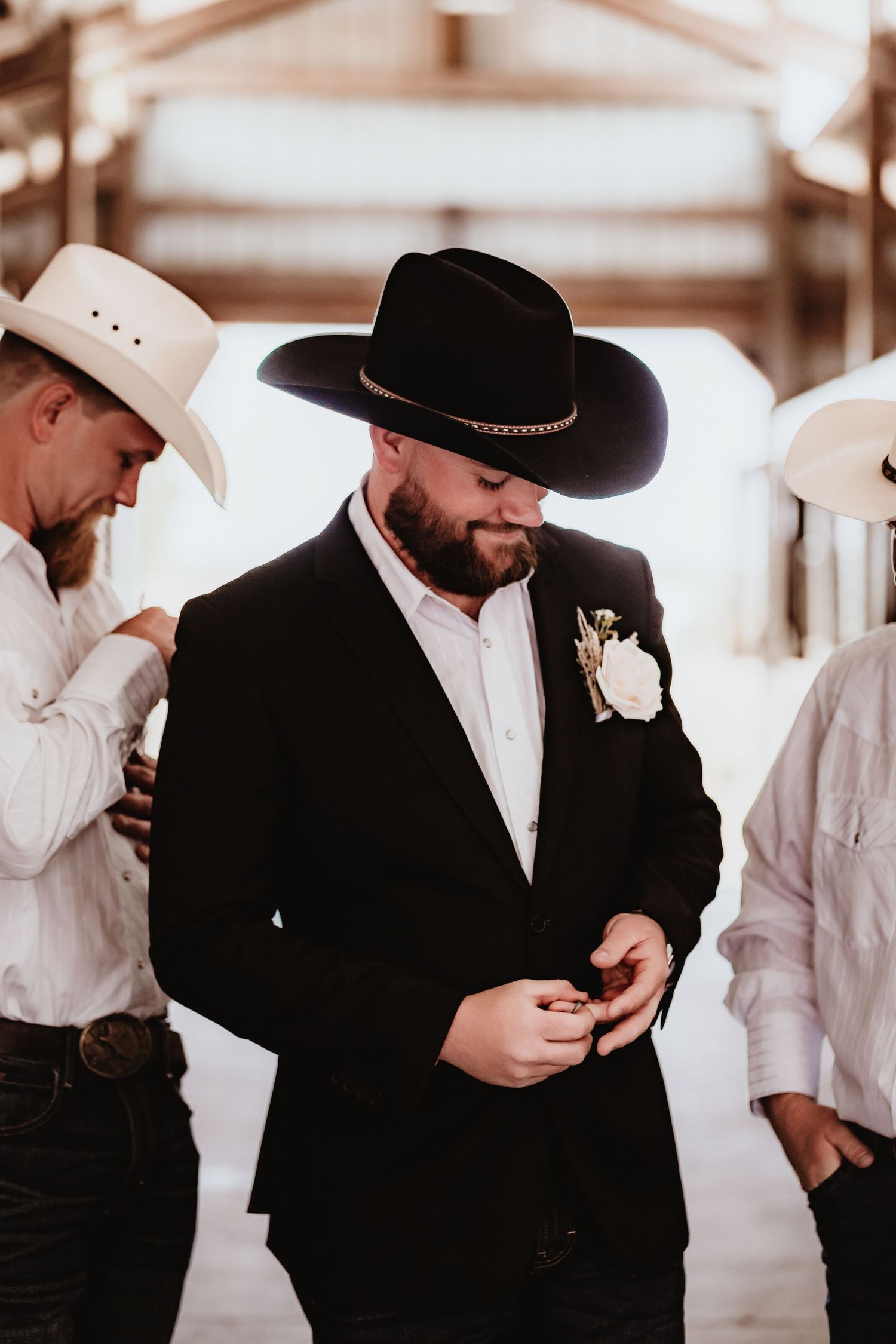 Bearded groom in black cowboy hat and suit adjusts cufflinks assisted by groomsmen in barn.