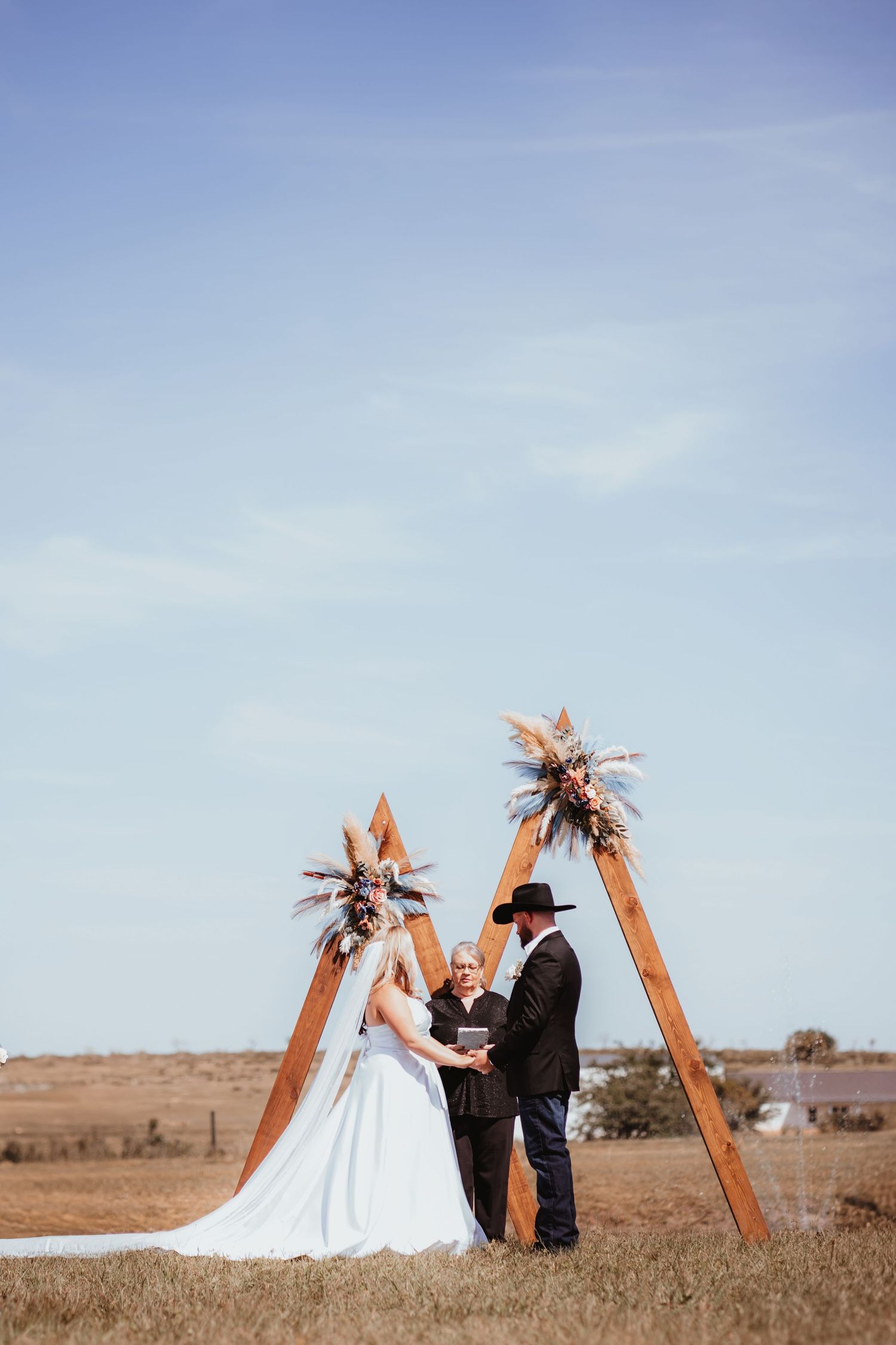 Bride and groom exchange vows under a rustic wooden triangular arch adorned with dried florals in an open field.