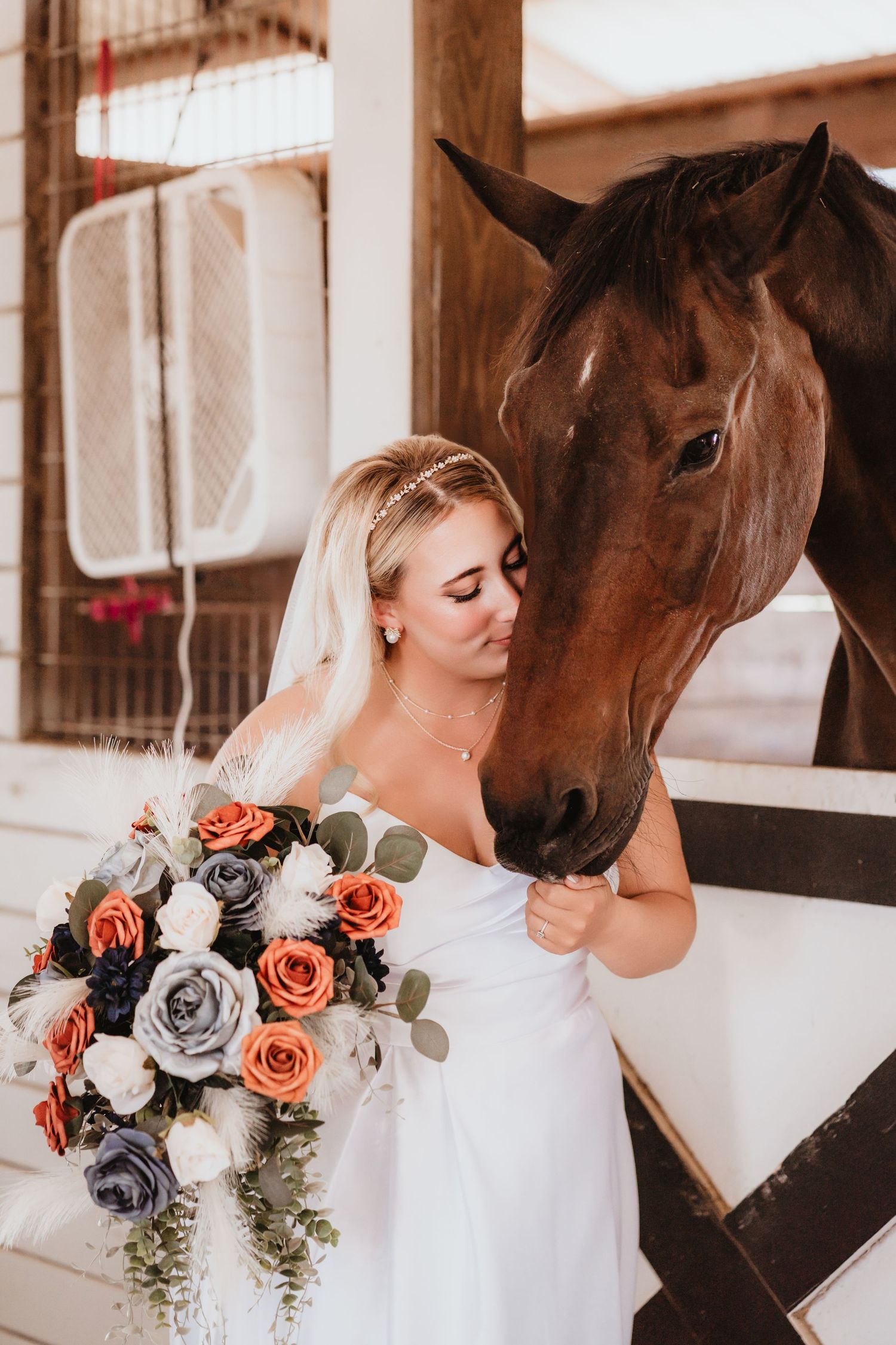 Bride in white gown holding colorful bouquet kisses a brown horse in a rustic stable.