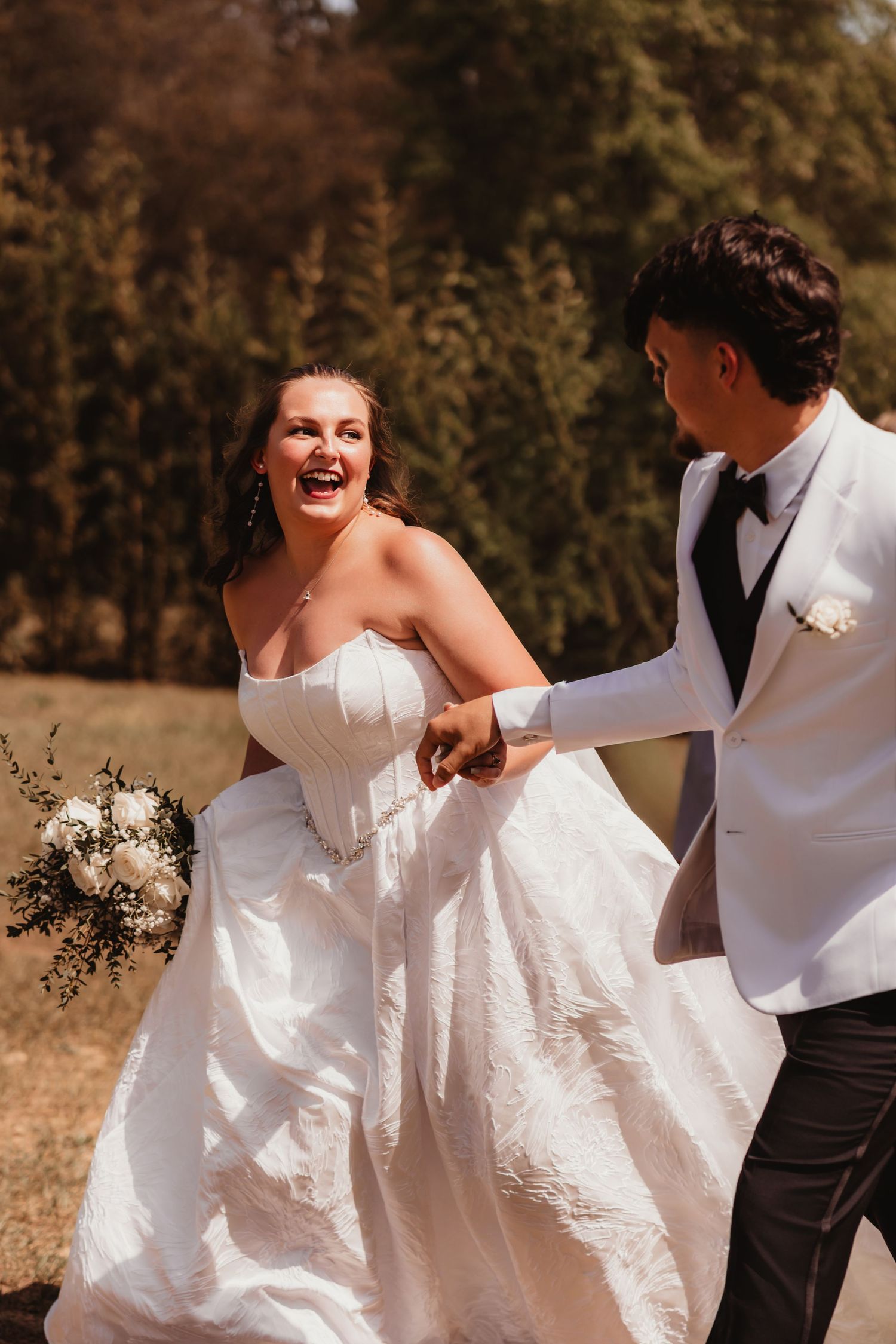 Laughing bride in strapless white gown holding bouquet while groom in white jacket leads her outdoors.