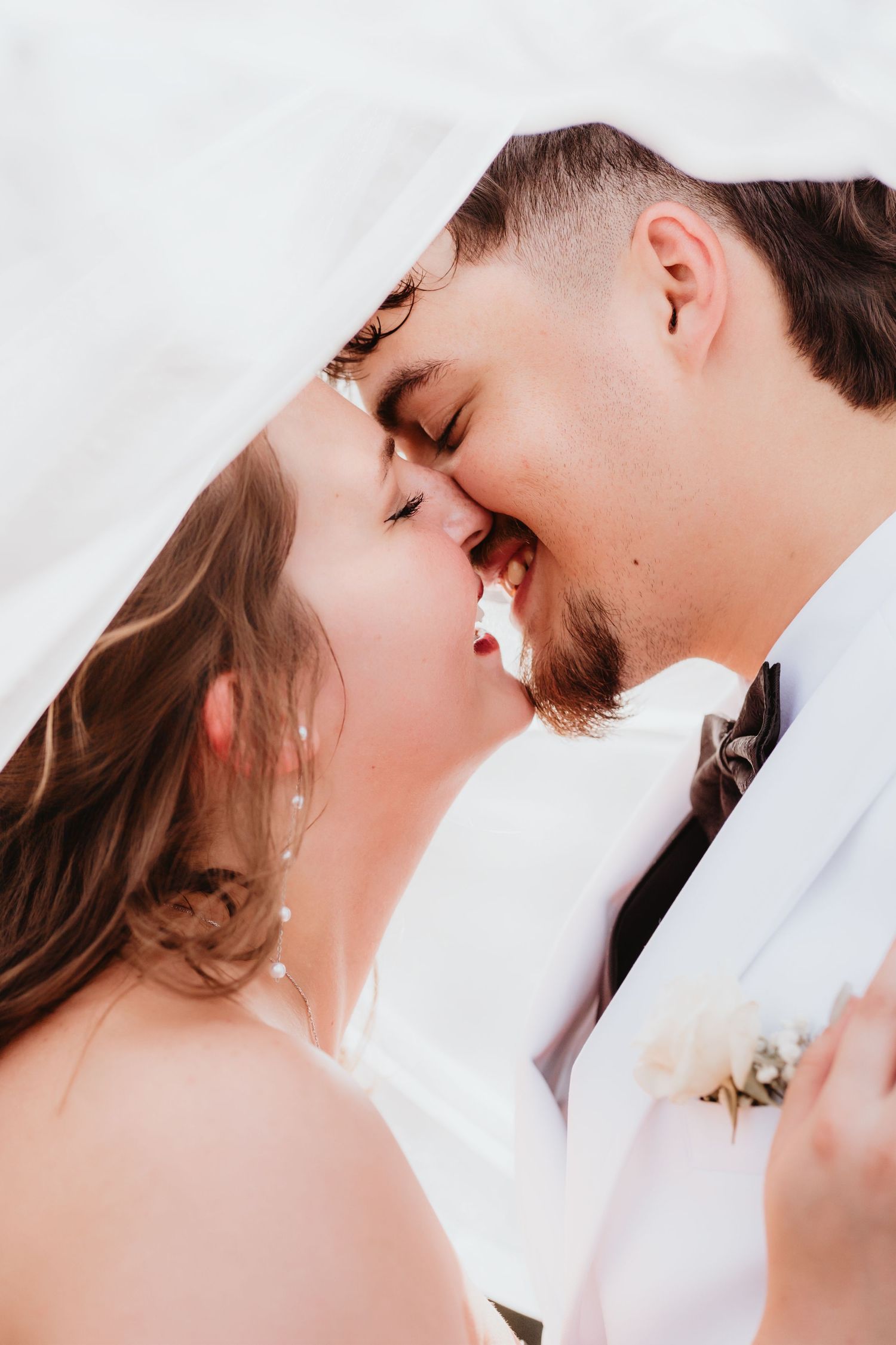Romantic close-up of bride and groom almost kissing under flowing white veil on wedding day.