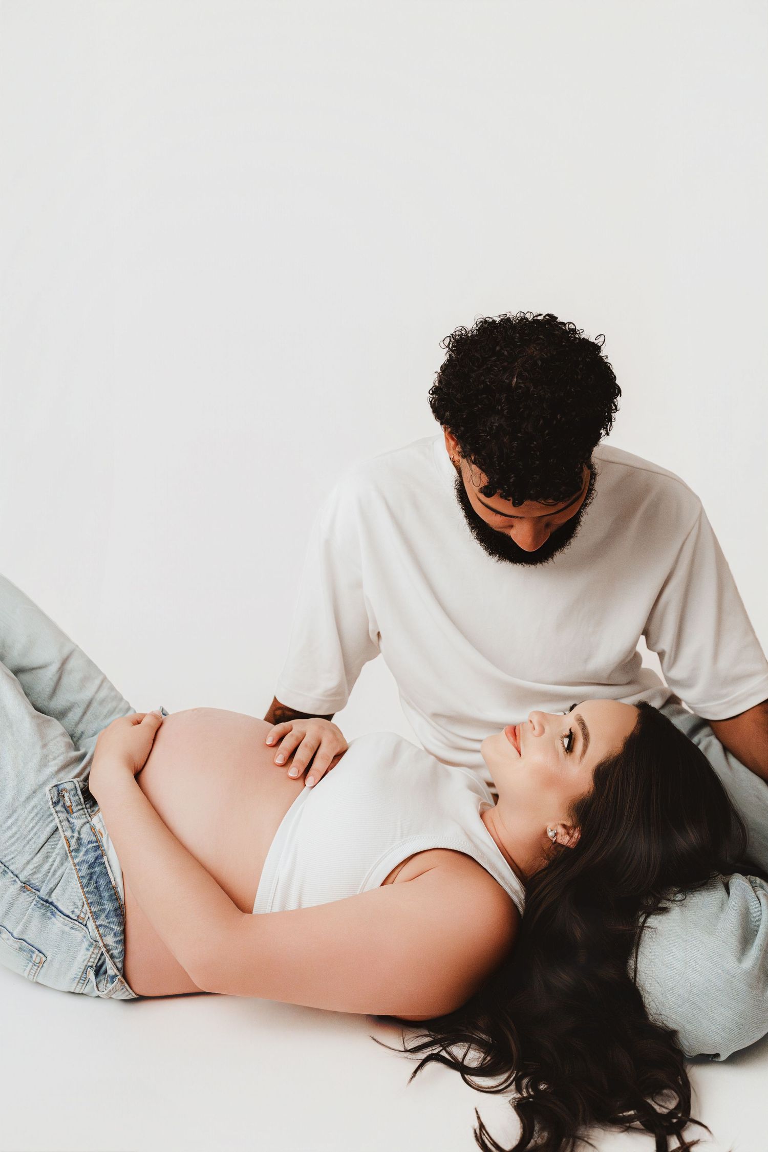 Pregnant woman lying down smiling up at partner who gently touches her bare baby bump.