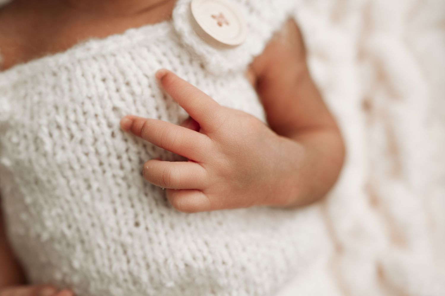 Close-up of newborn baby hand resting on white knit romper with button detail.