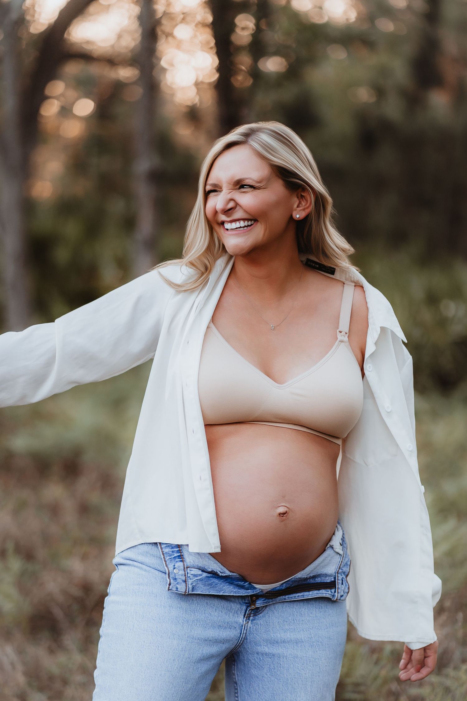 Joyful pregnant woman in white shirt and beige bralette laughing outdoors in forest.