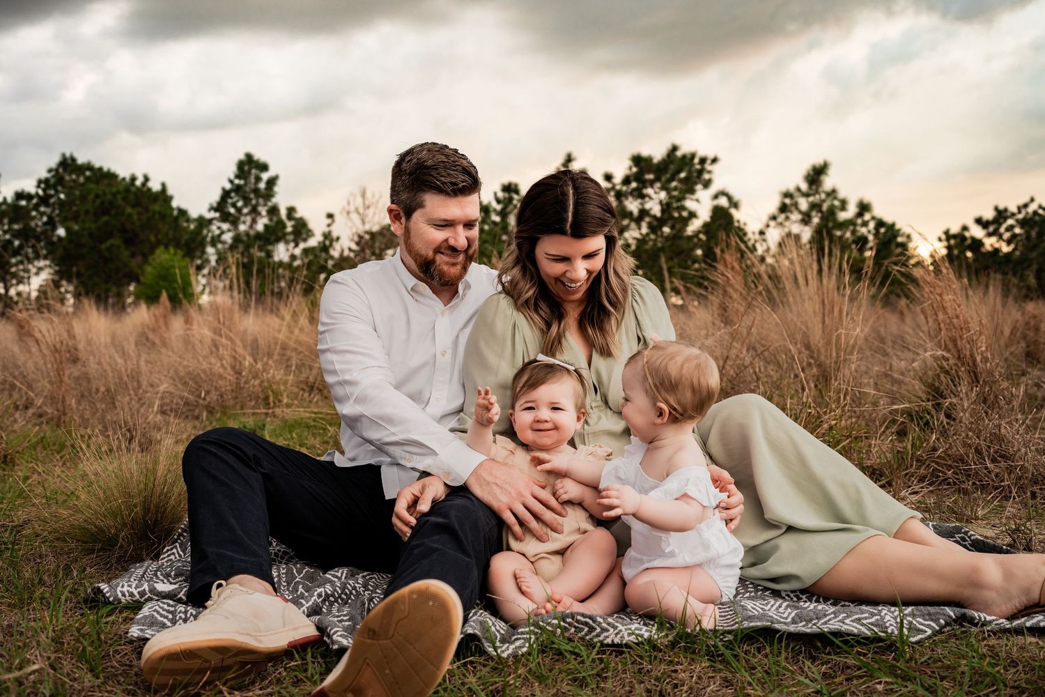 Happy family of four sitting on a blanket in a golden field at sunset, parents smiling at laughing toddler and baby.