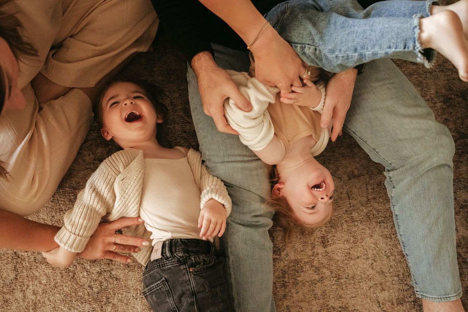 Two young children laughing joyfully while lying on a carpet with parents beside them at home.
