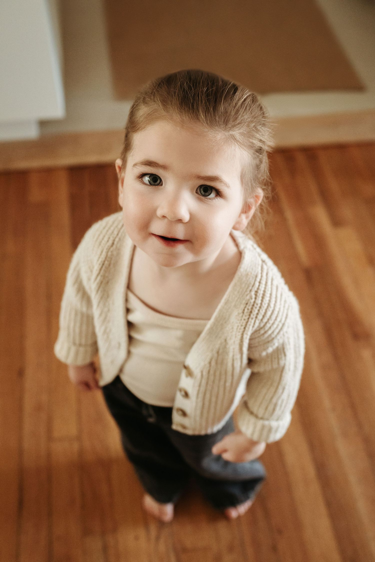 Cute toddler girl in beige cardigan and jeans looking up at camera while standing on hardwood floor indoors.