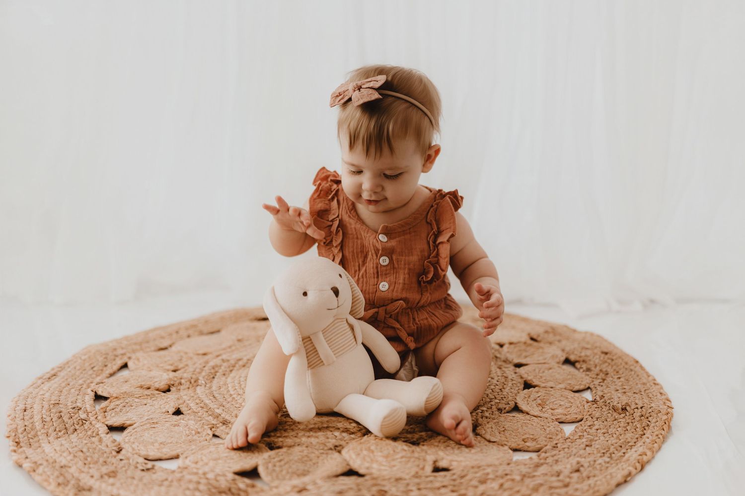 Baby girl in rust romper waving hand near stuffed bunny on woven round rug.