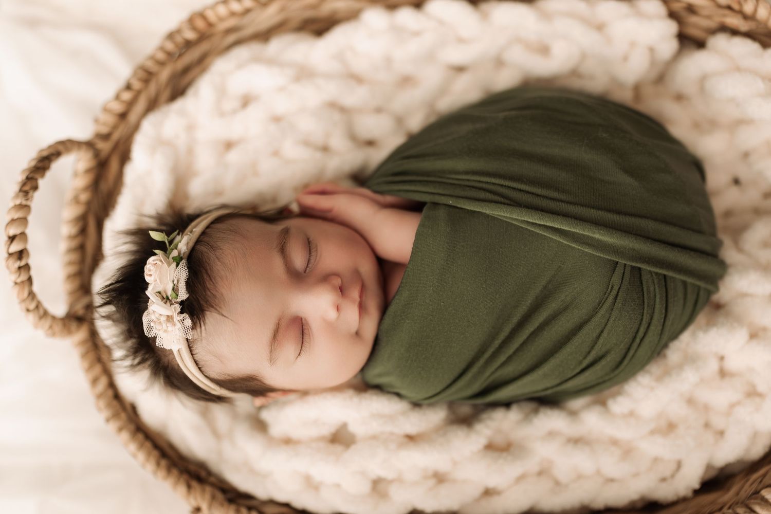 Close-up of newborn baby wrapped in green swaddle with floral headband resting in wicker basket.