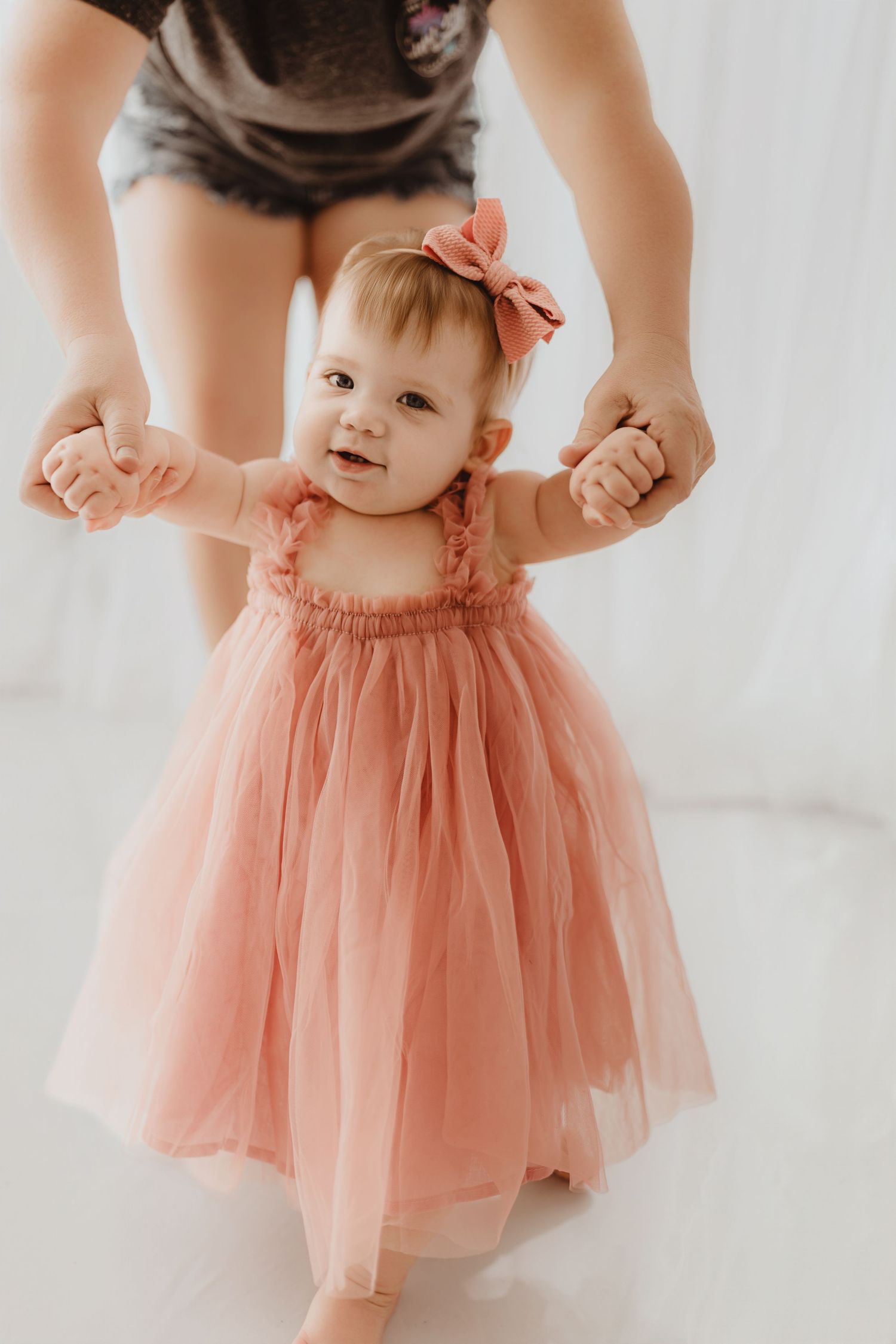 A baby girl in a pink tulle dress and bow takes first steps with adult support.