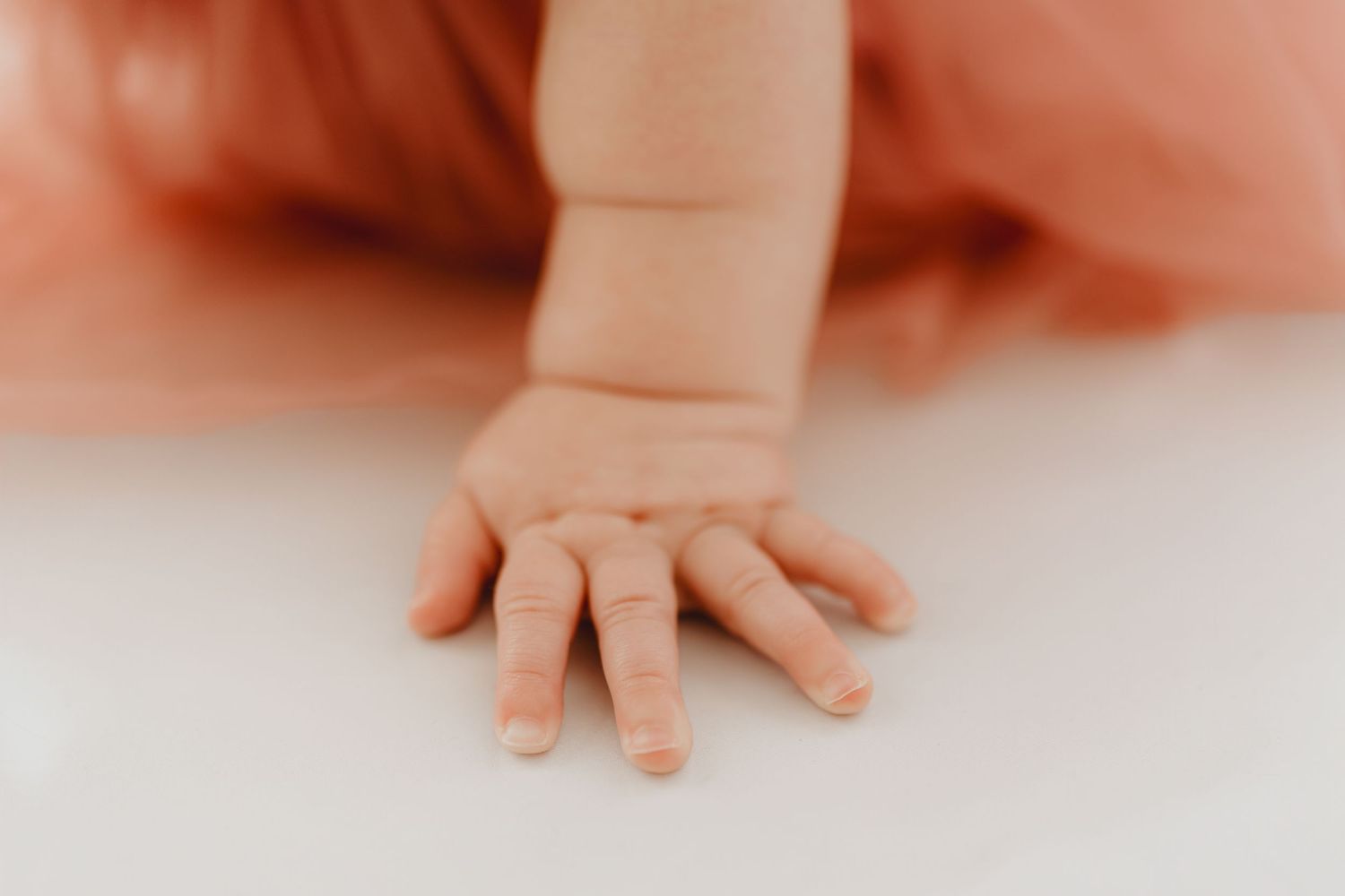 Delicate baby fingers spread on white surface with blurred pink tulle dress background.