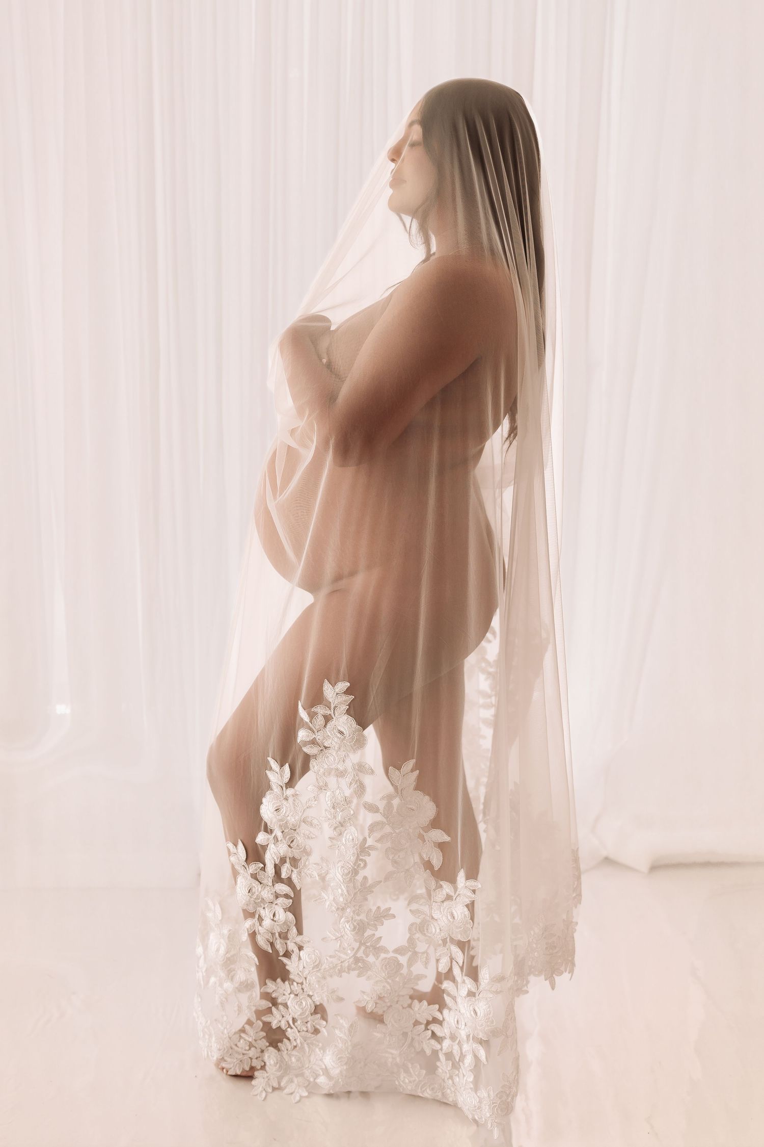 Pregnant woman draped in sheer white lace bridal veil in elegant studio setting.