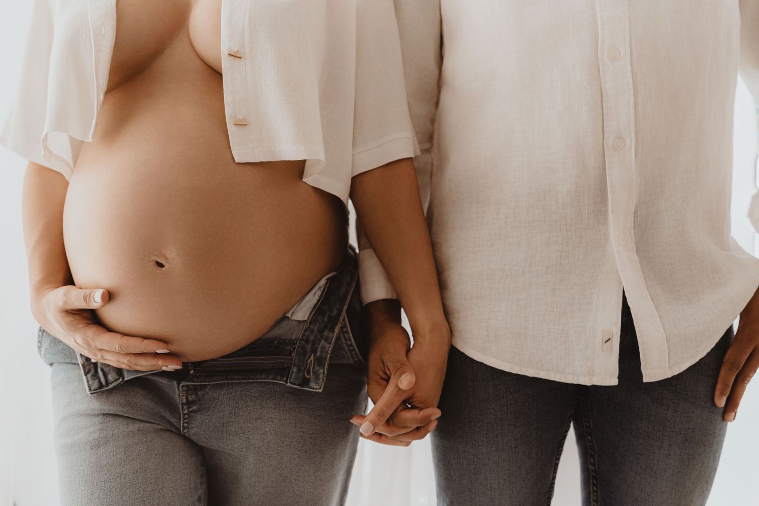 Pregnant woman in unbuttoned jeans holding hands with partner in white linen shirt.