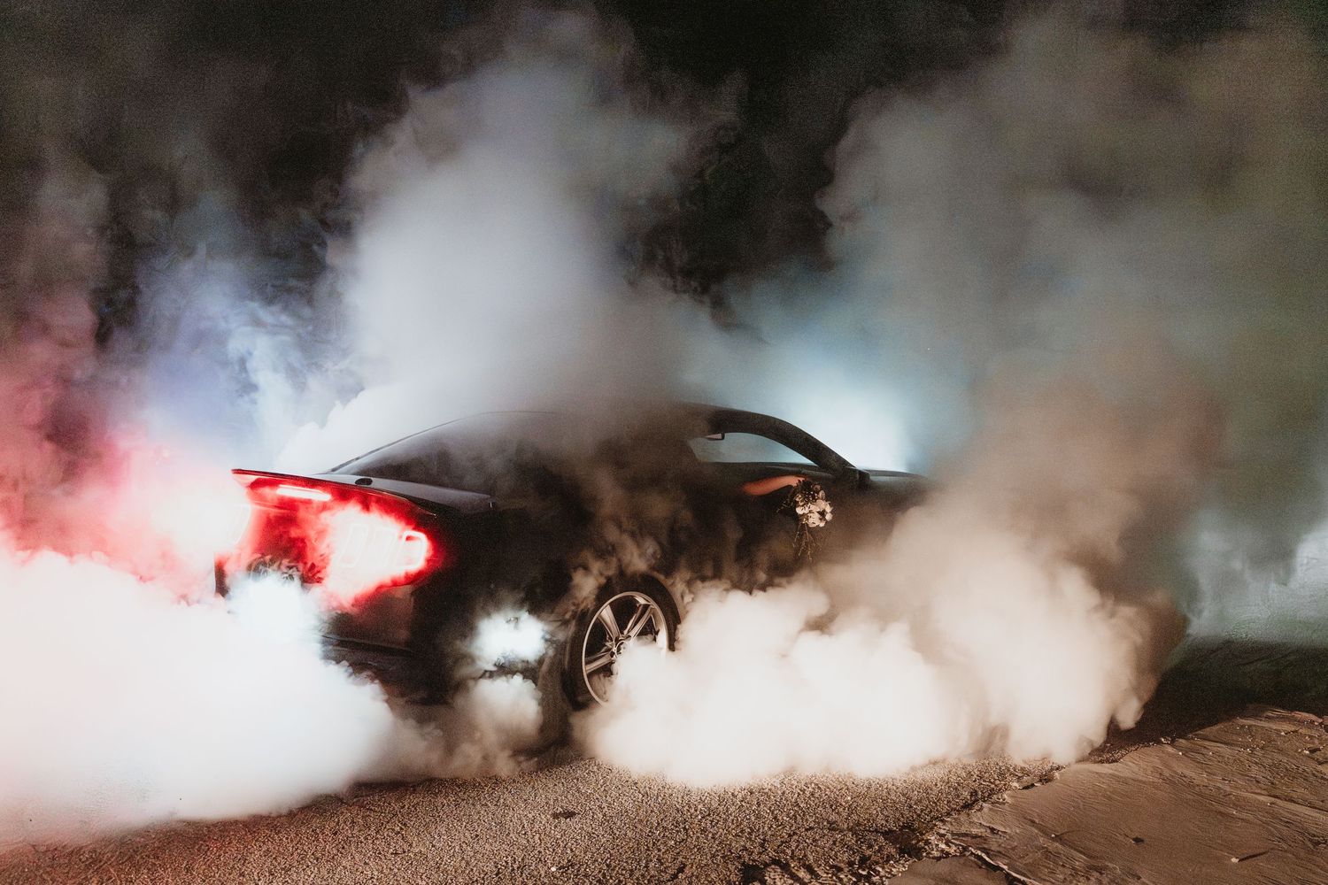 Sports car performing dramatic burnout surrounded by billowing white tire smoke at night on dark road.