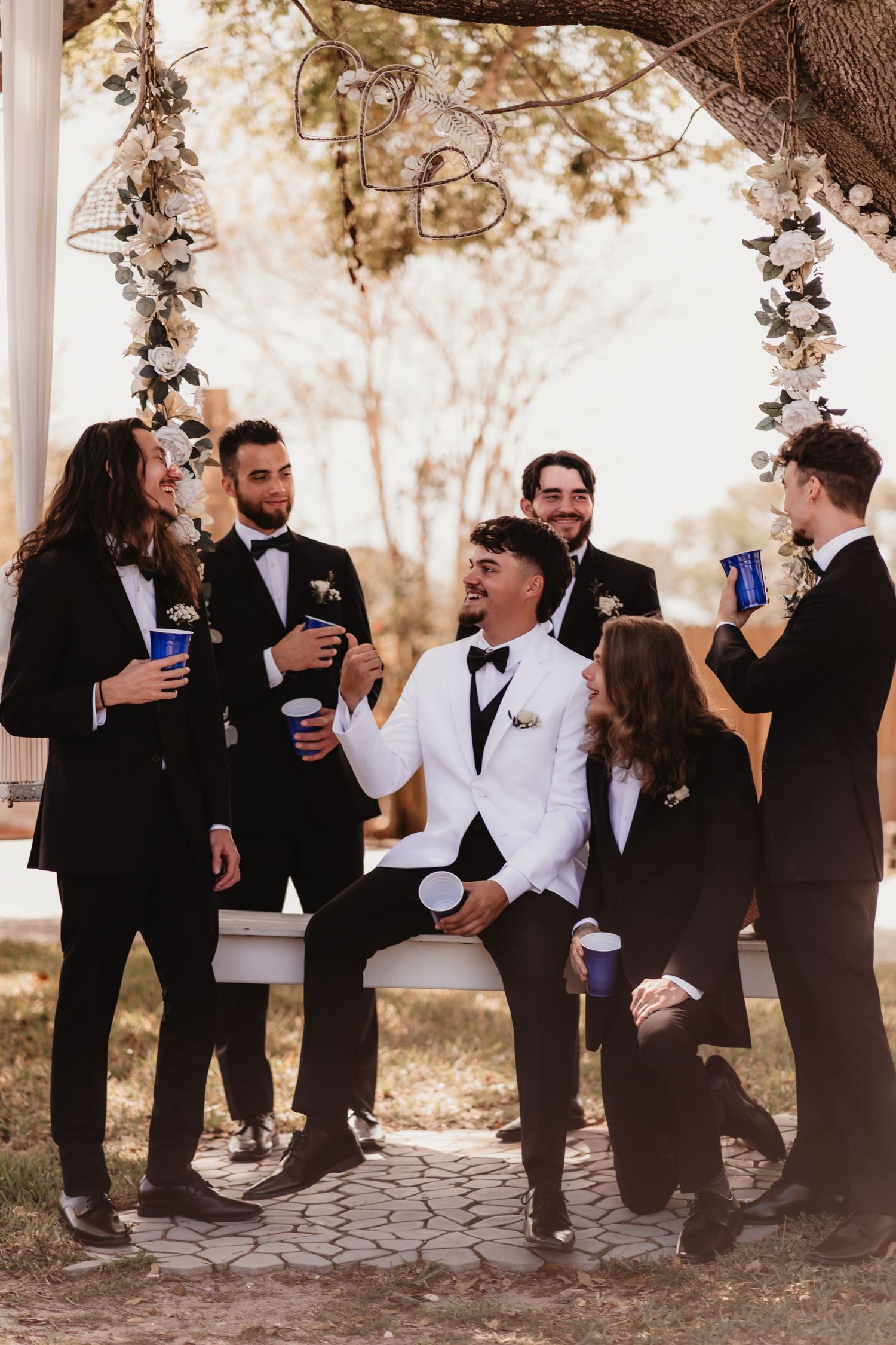 Groom in white tuxedo jacket celebrating with groomsmen in black suits under floral arch outdoors.