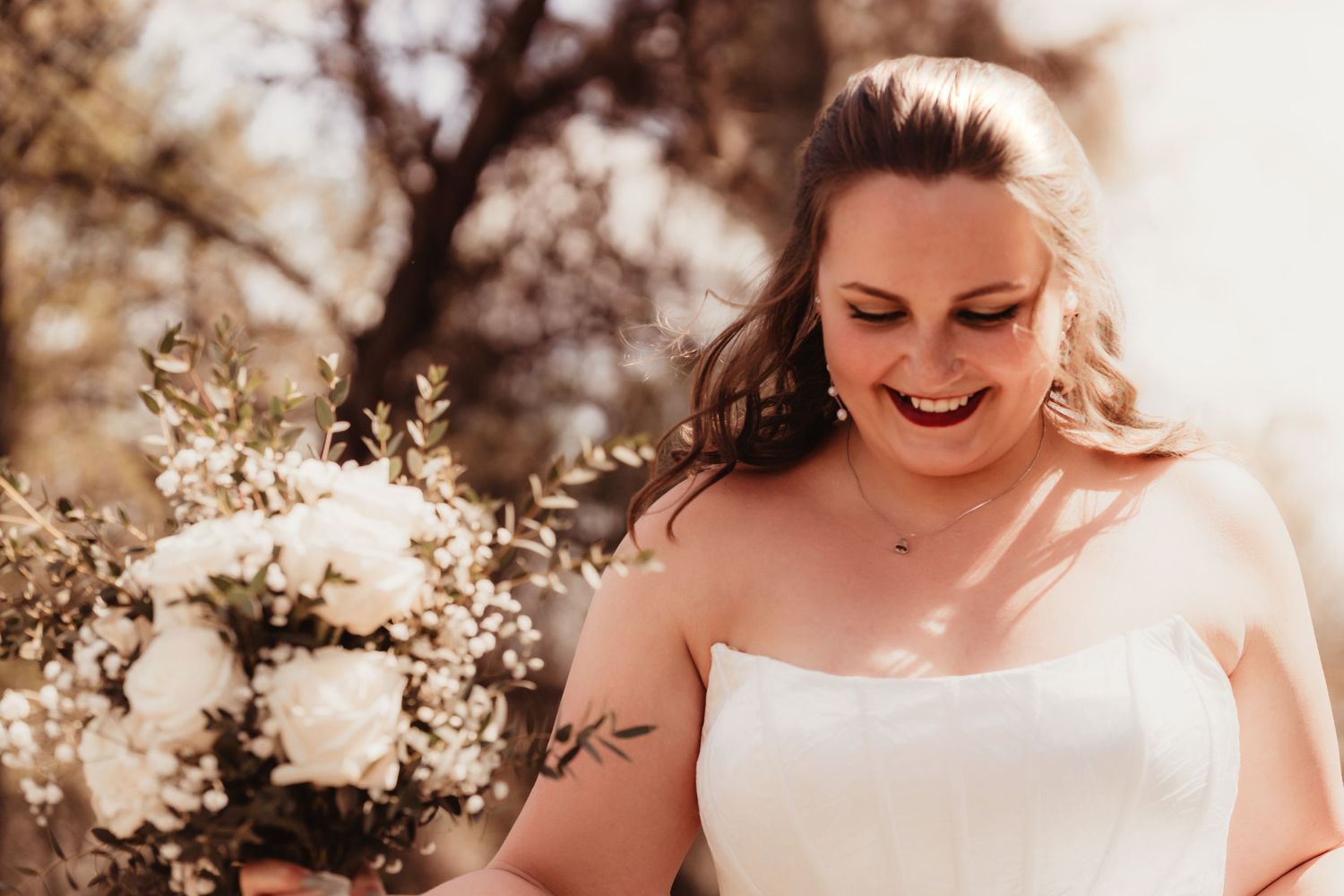 Radiant bride in strapless white gown smiling while holding white floral bouquet outdoors.