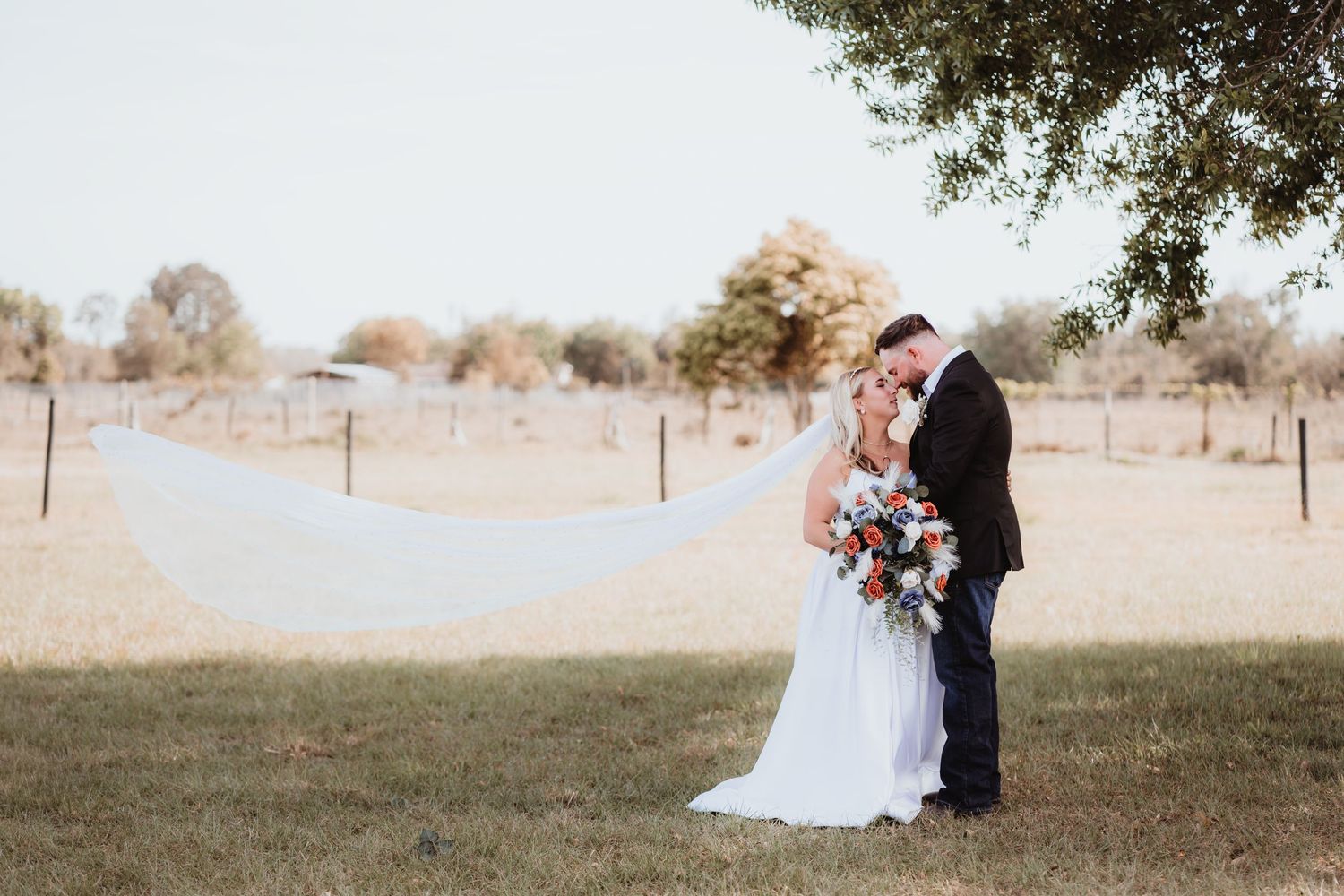 Bride and groom share a romantic moment under a tree with veil flowing in the wind at a rustic outdoor wedding venue.