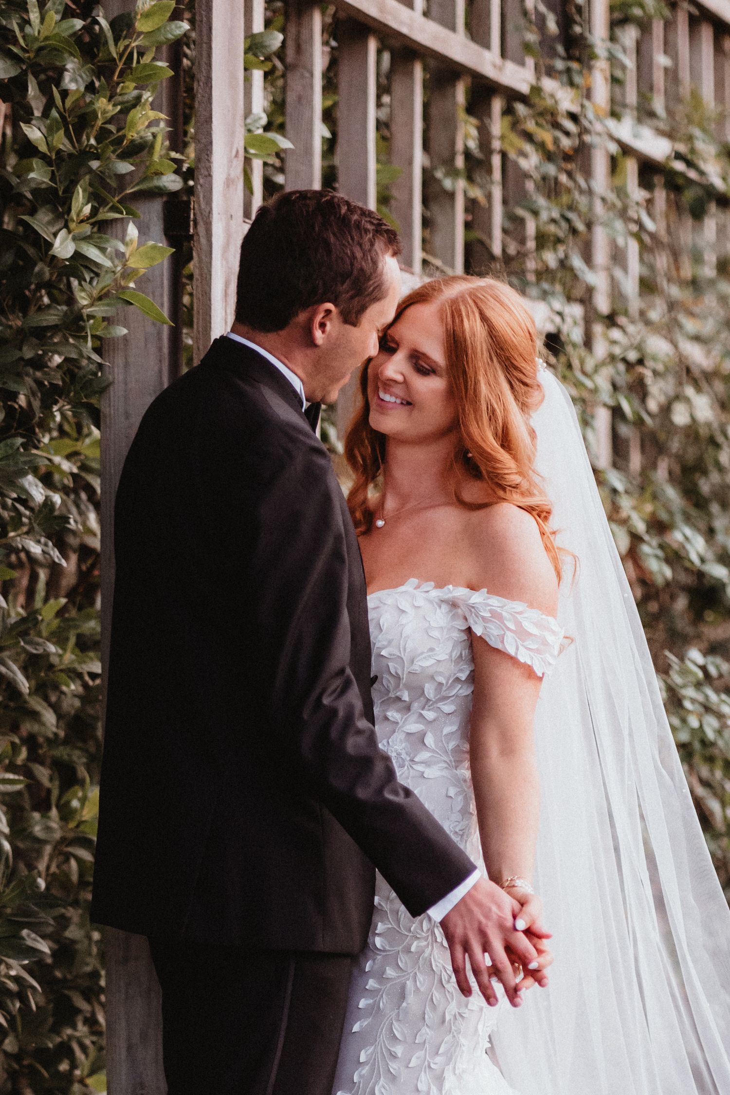 Bride and groom hold hands and share an intimate moment by garden trellis.