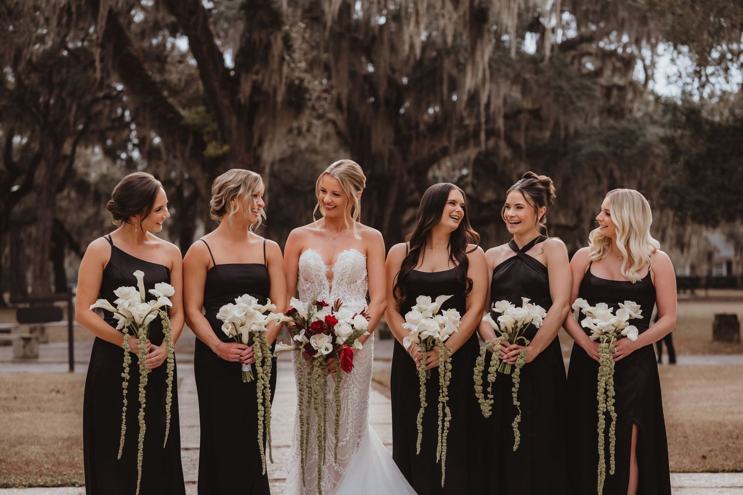 Bride in white gown smiles with five bridesmaids wearing black dresses holding white floral bouquets under oak trees.
