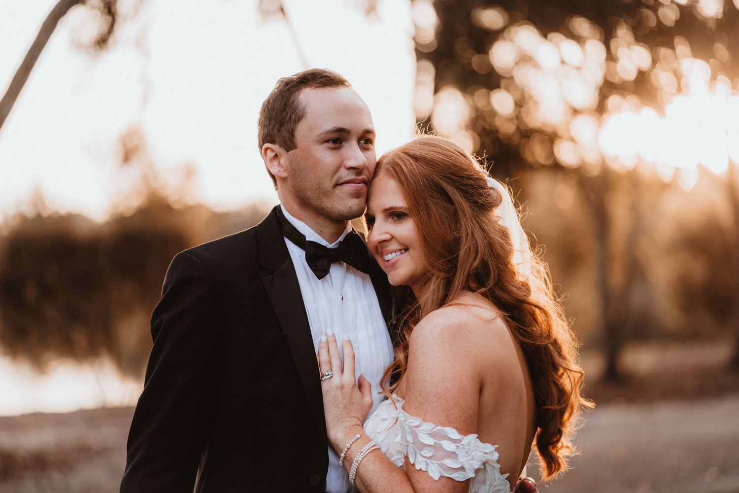 Bride with red hair leans into groom in black tuxedo during warm golden hour outdoor wedding portrait.