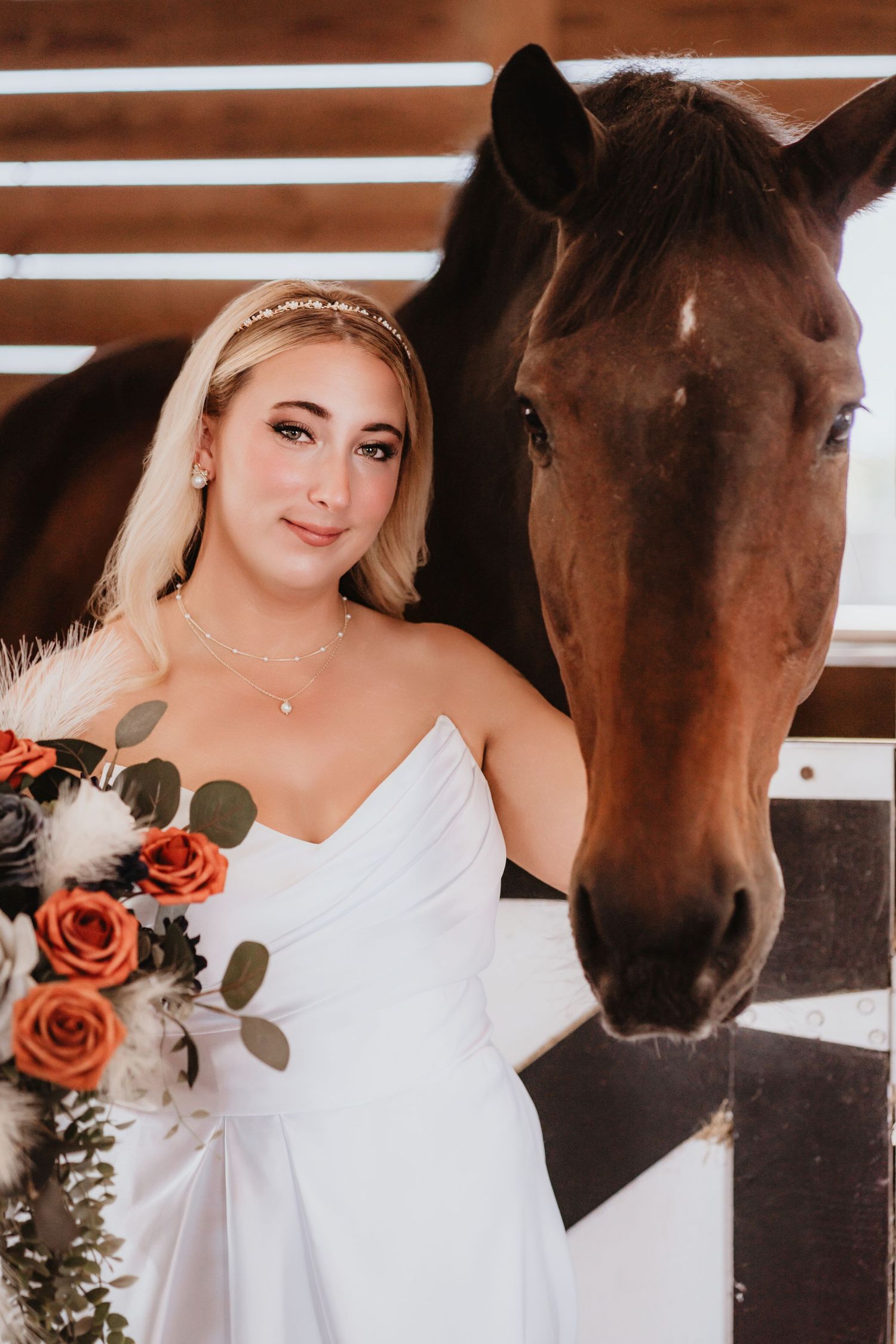 Elegant bride in white gown poses beside a brown horse in a sunlit barn stable.