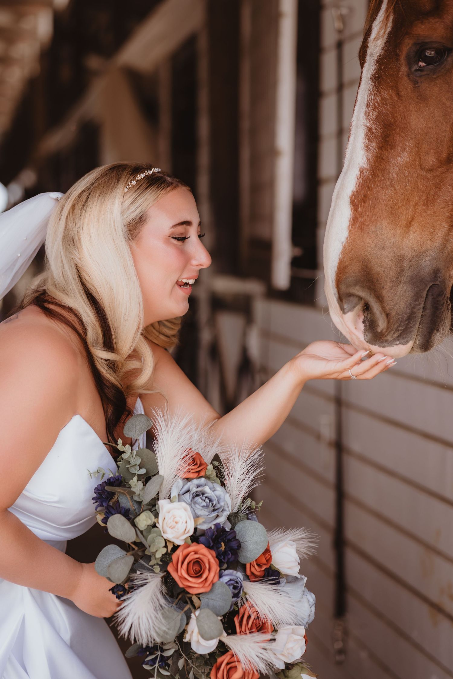 Smiling bride in white dress feeds a chestnut horse while holding a colorful wedding bouquet.
