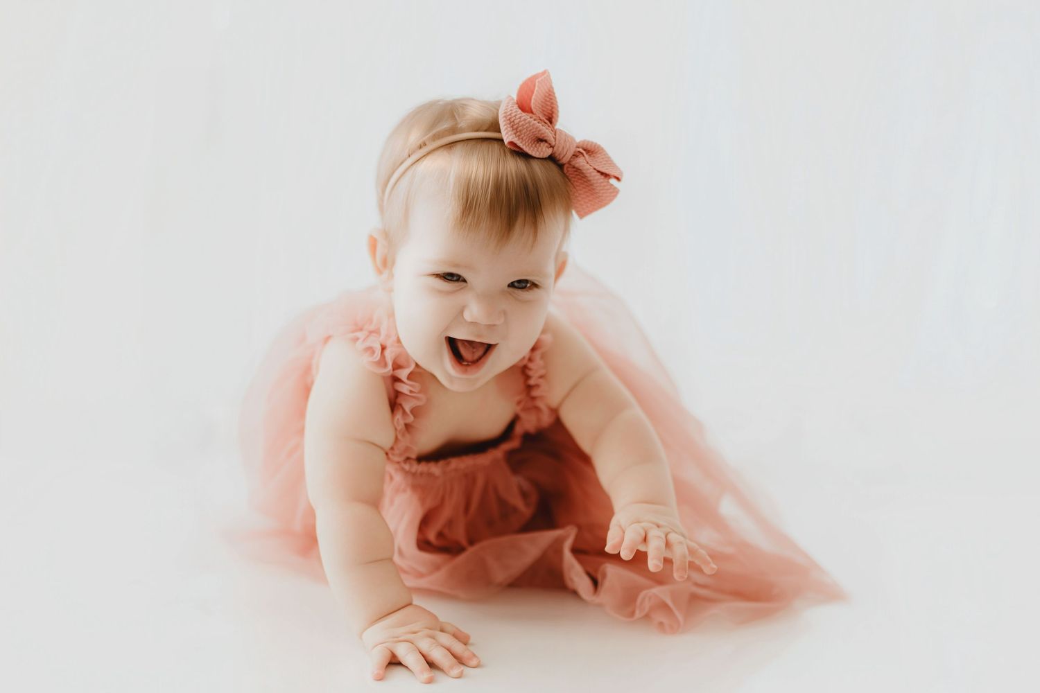 Happy laughing baby girl in pink ruffle dress and bow headband crawling on white background.