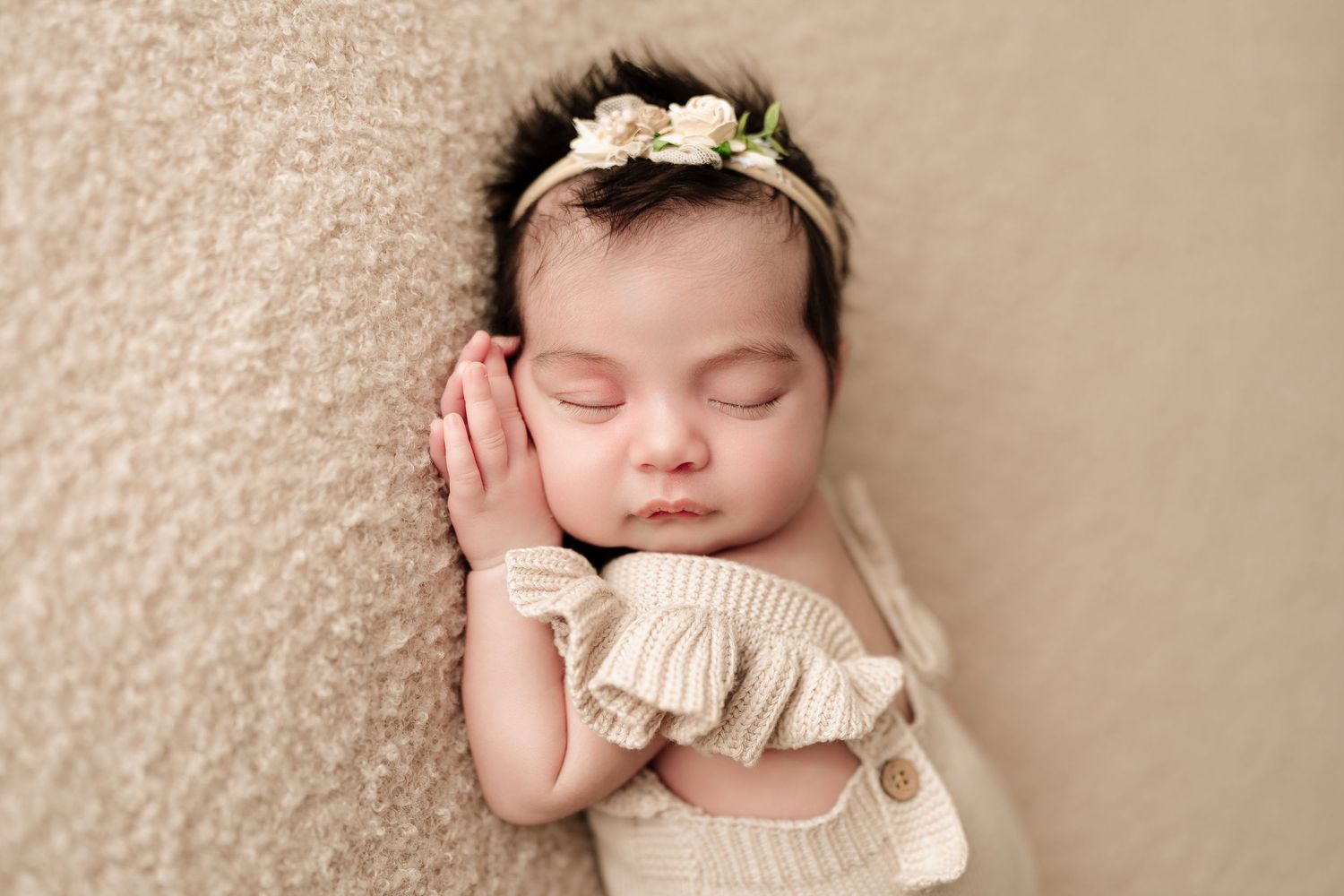 Adorable sleeping newborn baby girl in cream knit romper and floral headband posed on beige backdrop.