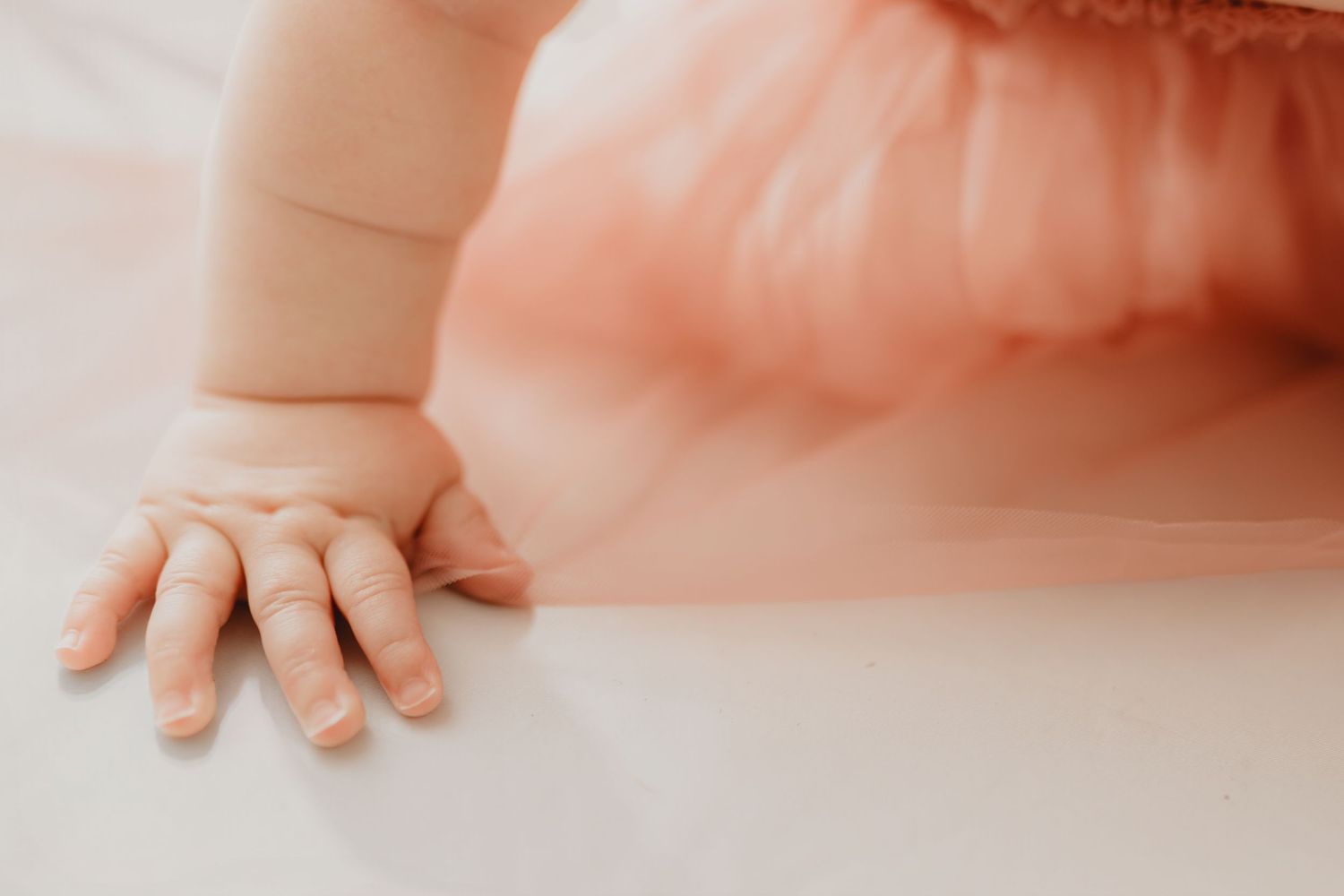 Close-up of a baby girl's tiny hand on white floor in pink tulle dress.
