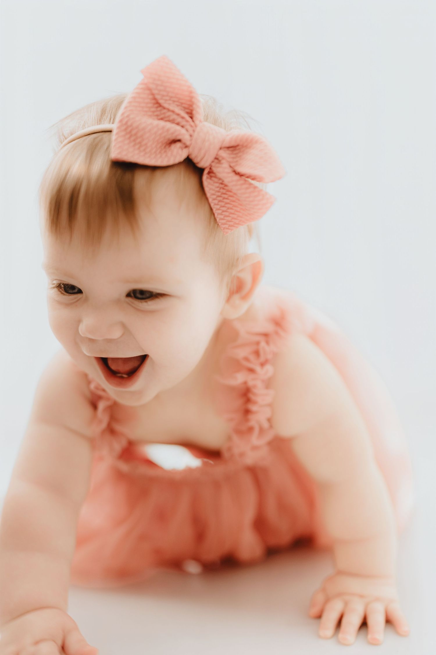 Giggling baby girl in coral ruffled romper with pink bow headband during white background photo session.