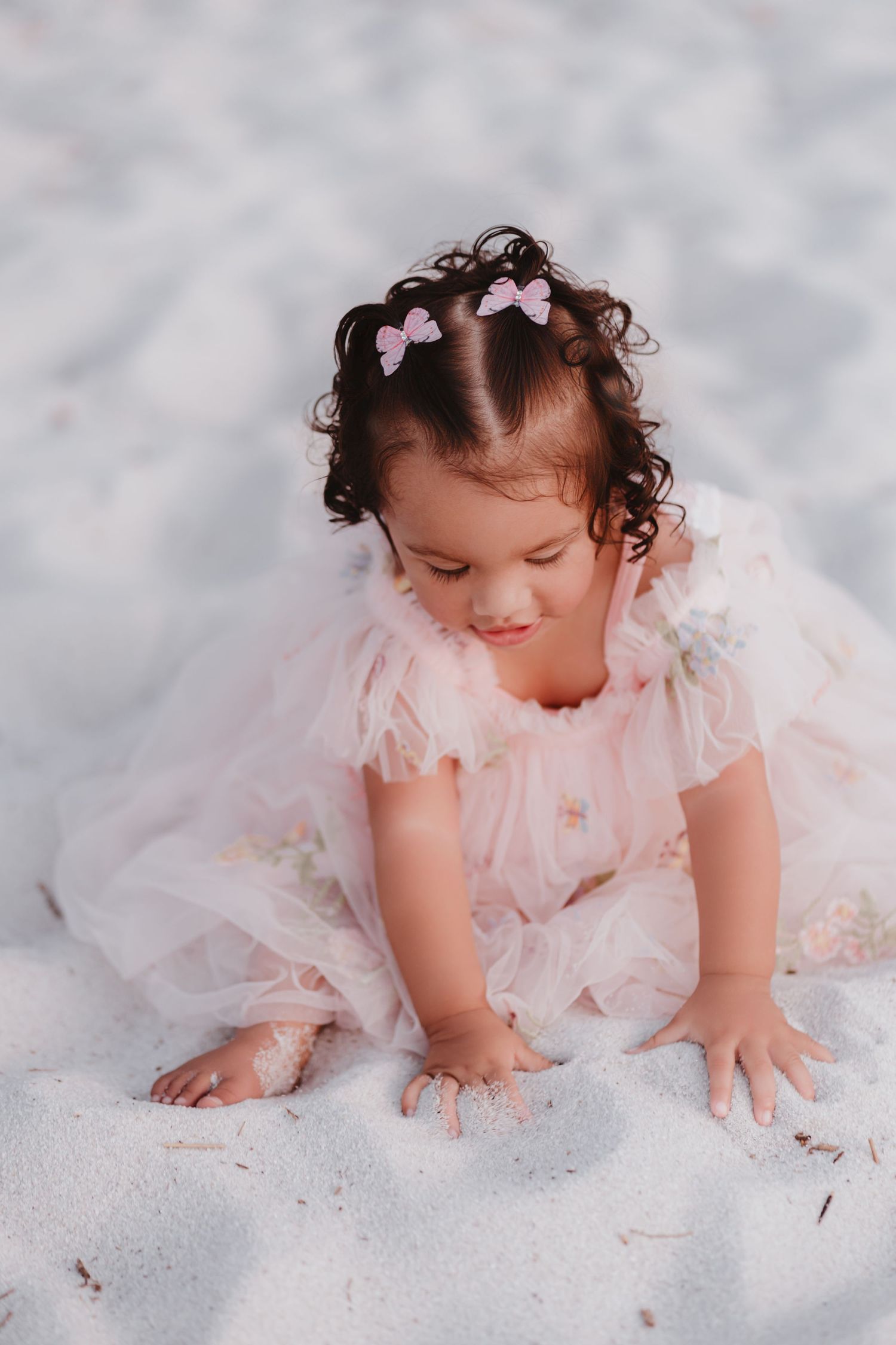 Toddler girl in pink tulle dress with bow hair clips playing in white sand at the beach.