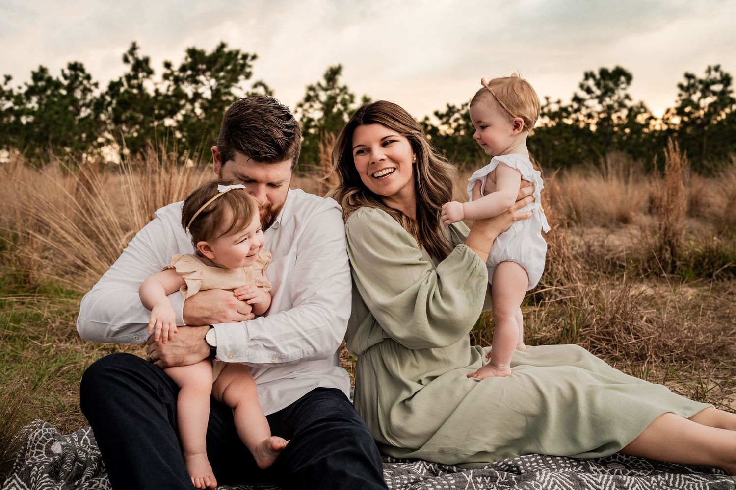 Laughing family of four with two babies sitting on a blanket in a warm golden sunset field setting.