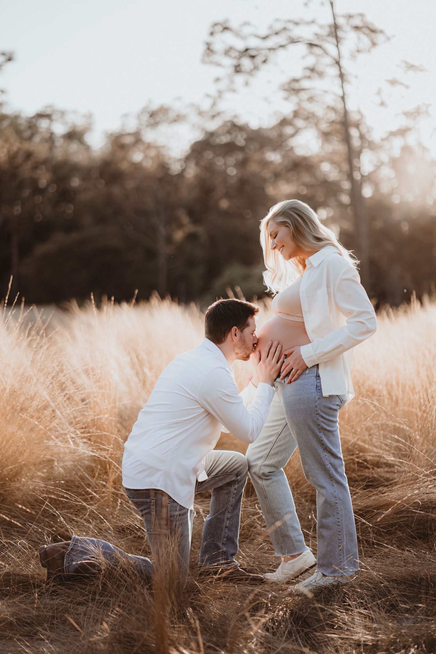 Man kneeling to kiss pregnant partner's belly in sun-drenched golden grass field.