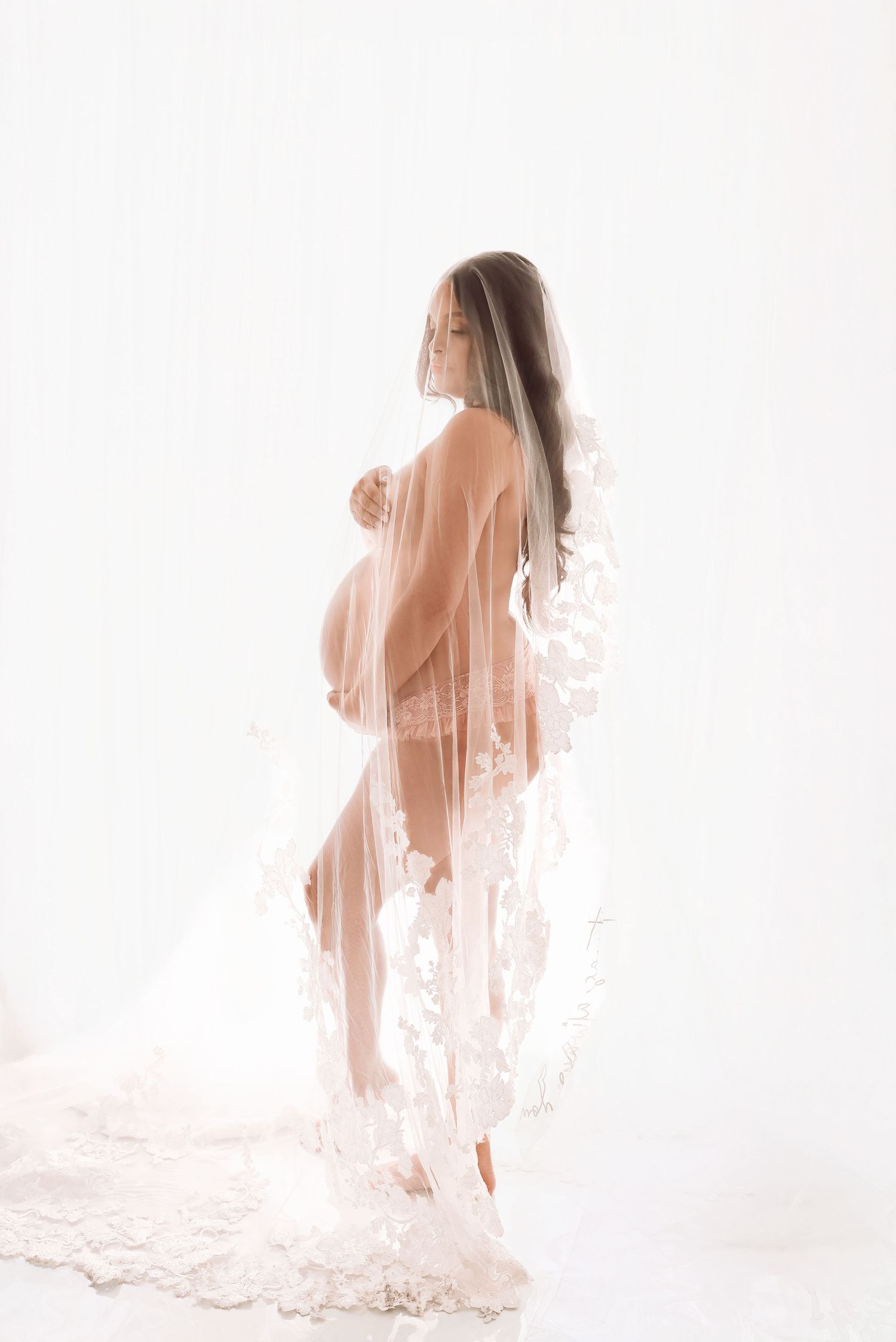 Elegant pregnant woman posed in sheer lace veil, showcasing her baby bump in a soft, ethereal maternity portrait.