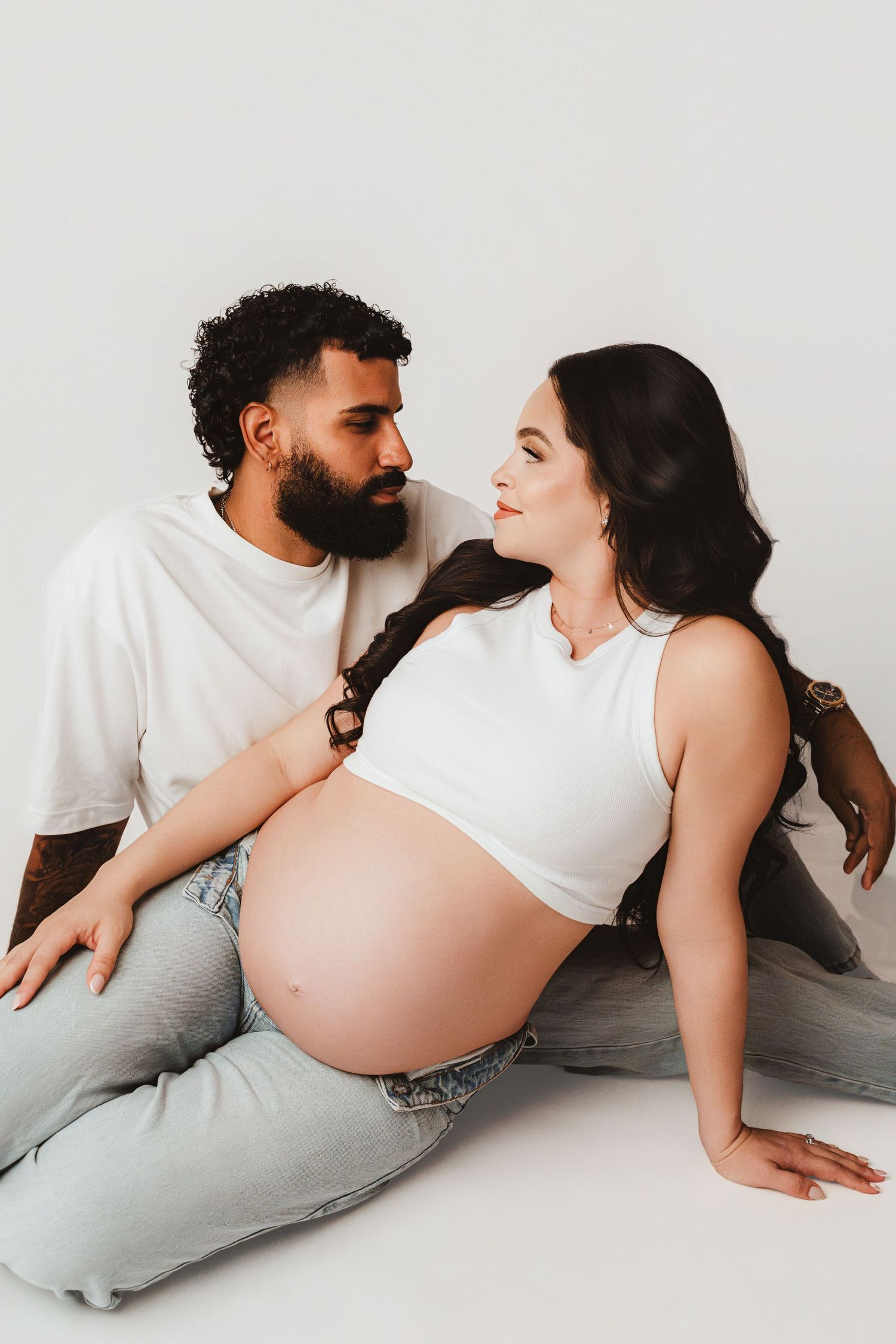 Pregnant woman in white crop top and jeans sitting with partner against white background.