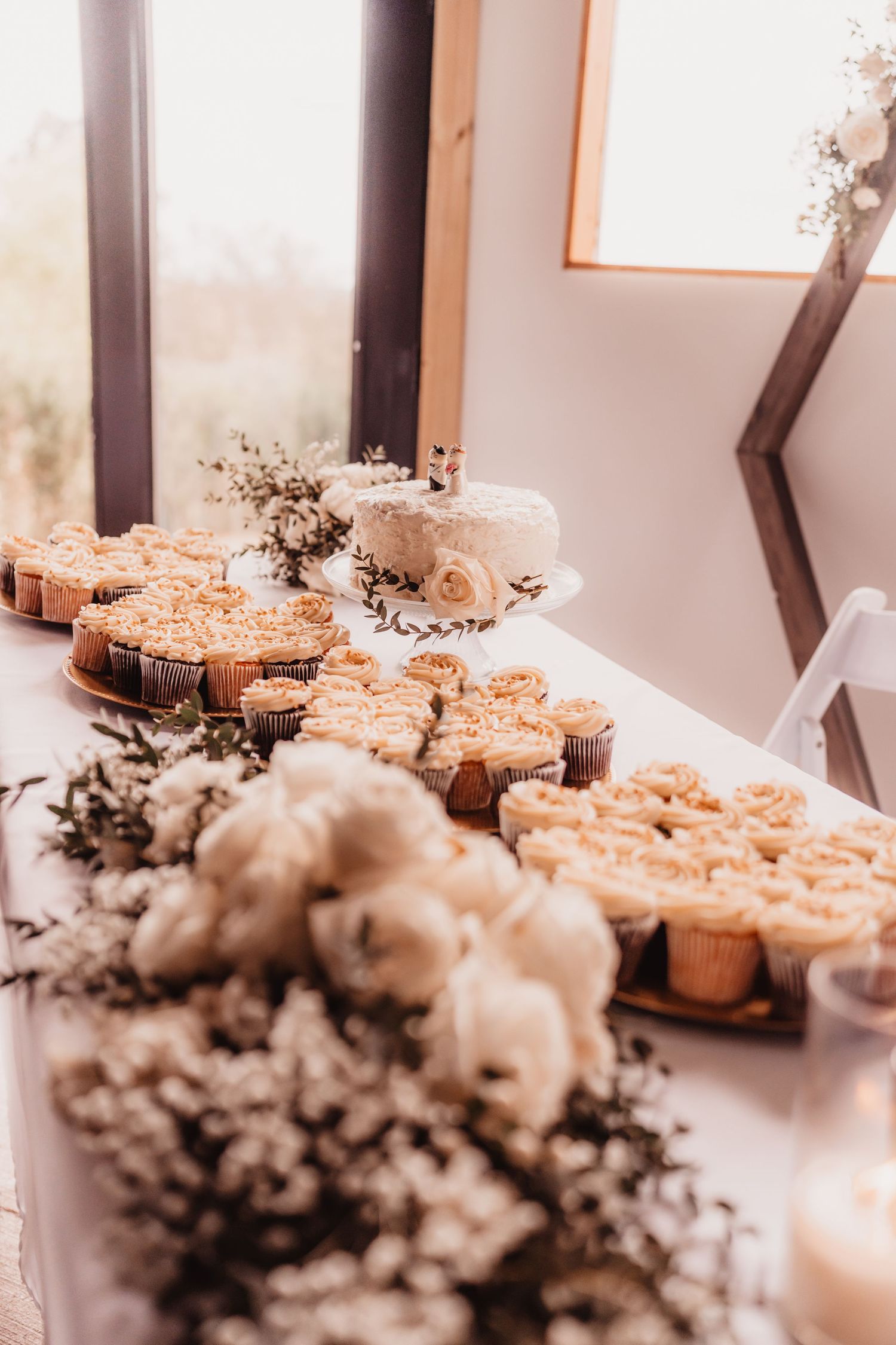 Wedding dessert table featuring a white cake, caramel cupcakes, and white floral arrangements.