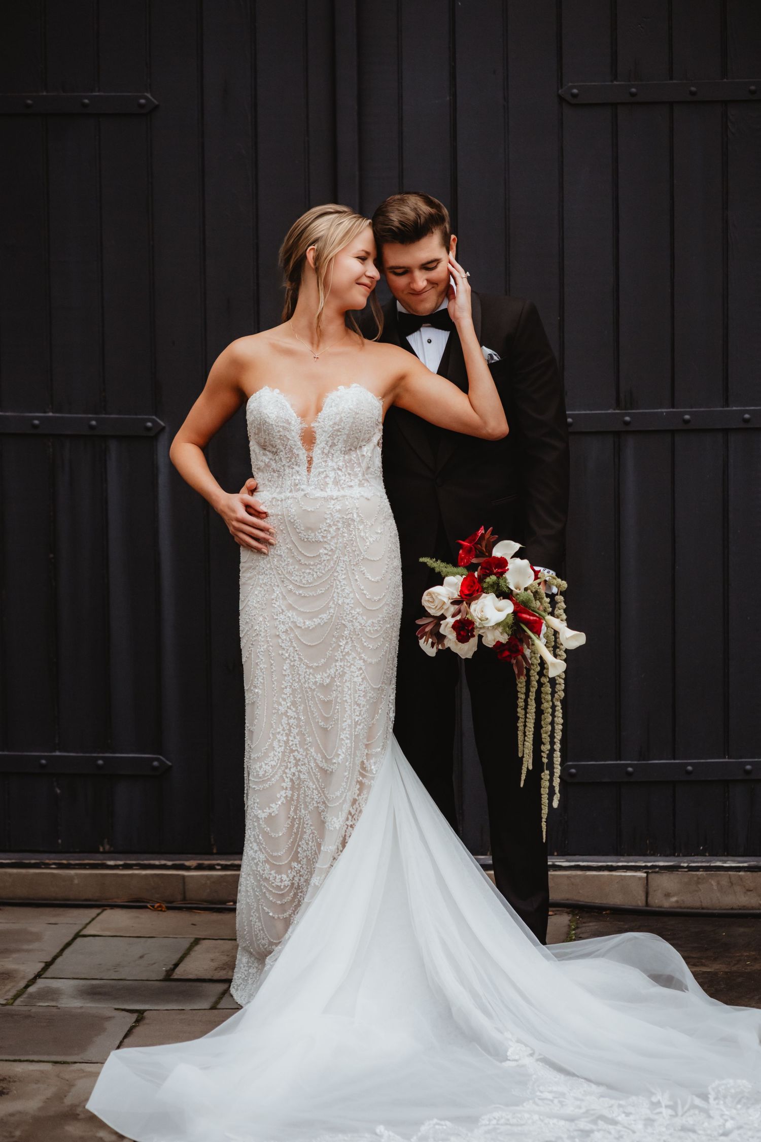 Bride in strapless beaded gown and groom in black tuxedo pose together holding a red and white bouquet.