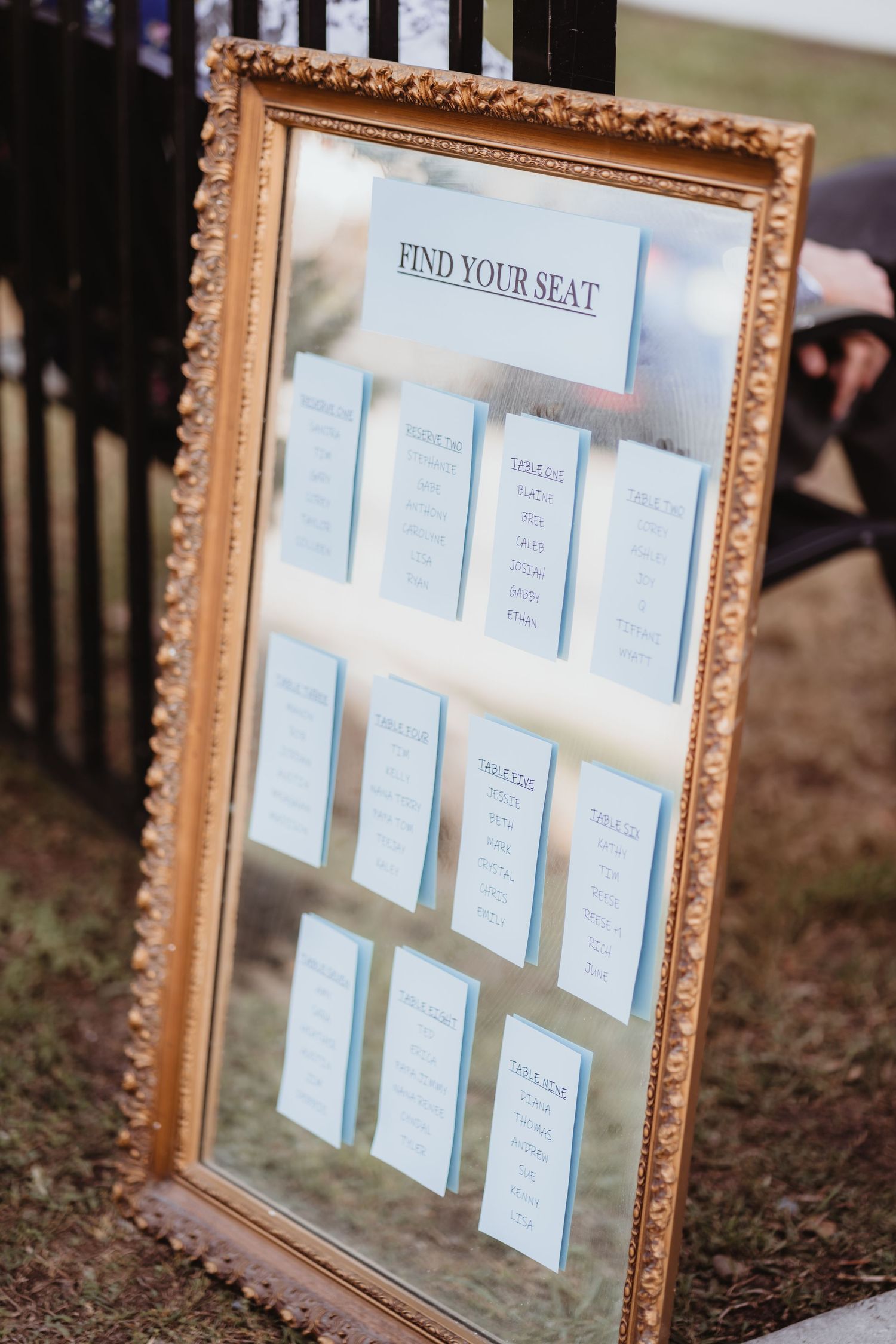 Gold-framed mirror seating chart displaying 'Find Your Seat' with blue card table assignments for a wedding reception.