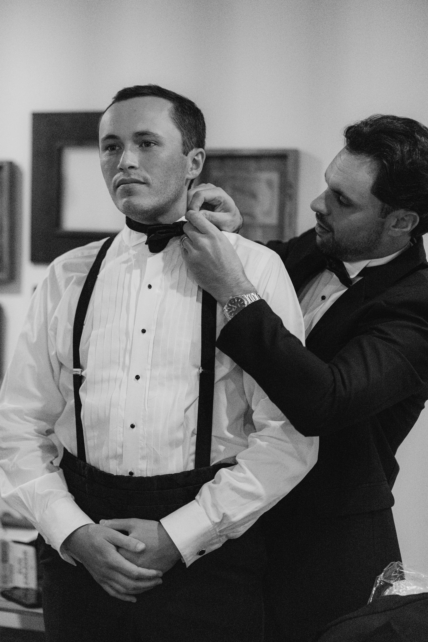 Black and white photo of groomsman helping groom adjust bow tie before wedding.