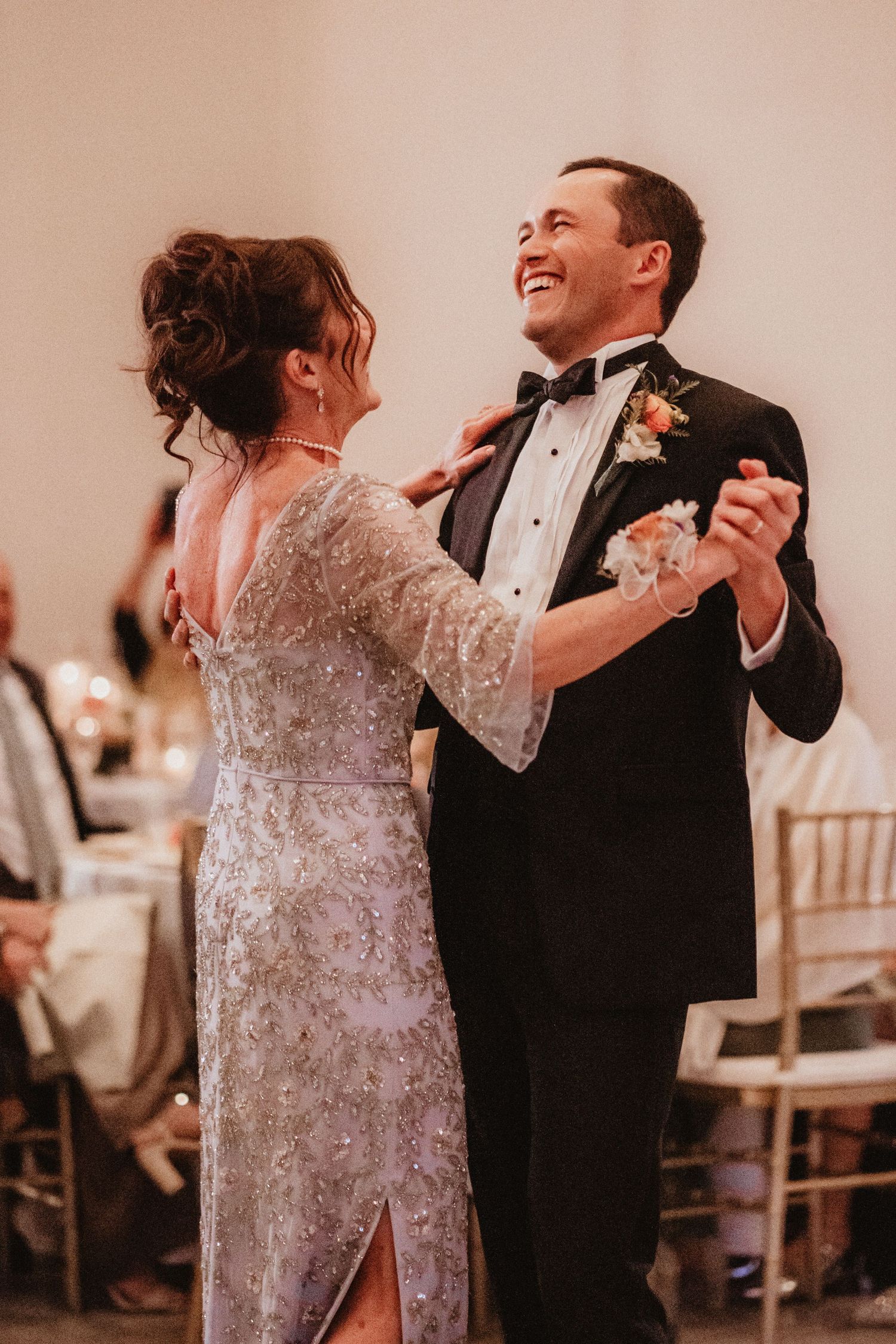Groom in black tuxedo shares a joyful mother-son dance with mom in silver beaded gown at wedding reception.