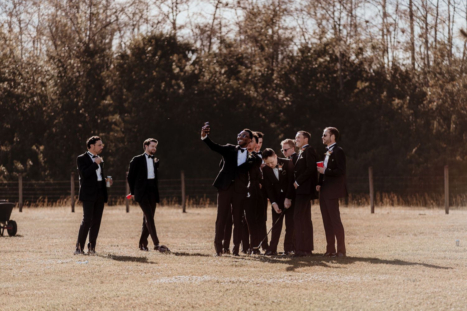 Wedding party group gathers and gestures on an open sunny field during pre-ceremony photos.