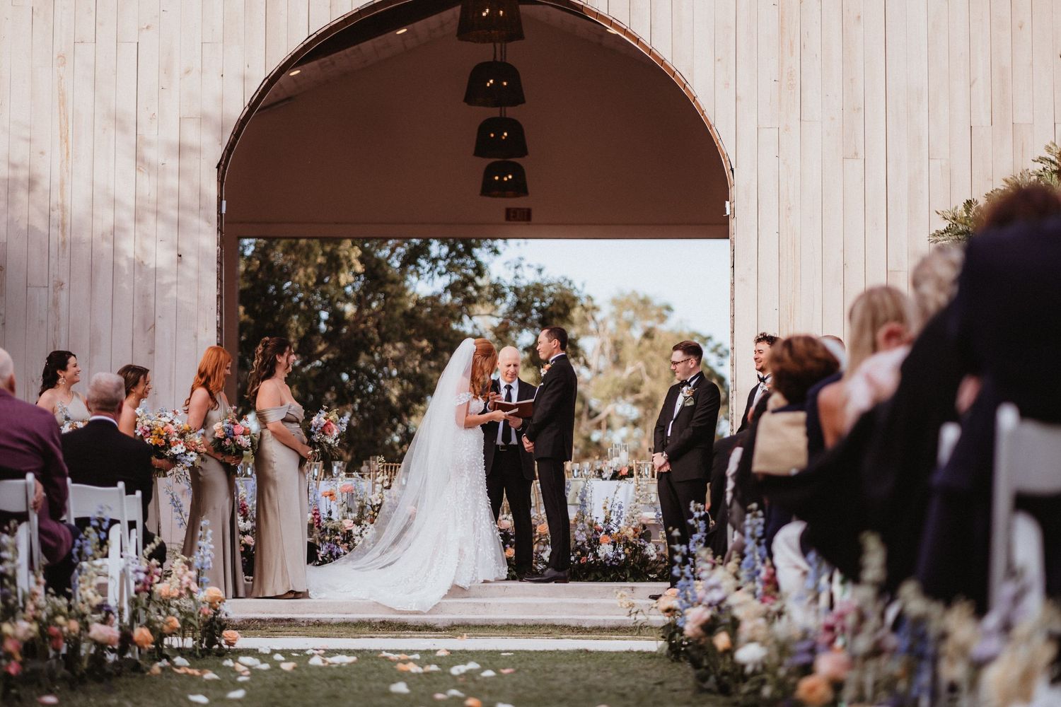 Wedding ceremony under rustic barn arch with bride and groom at altar surrounded by floral arrangements.