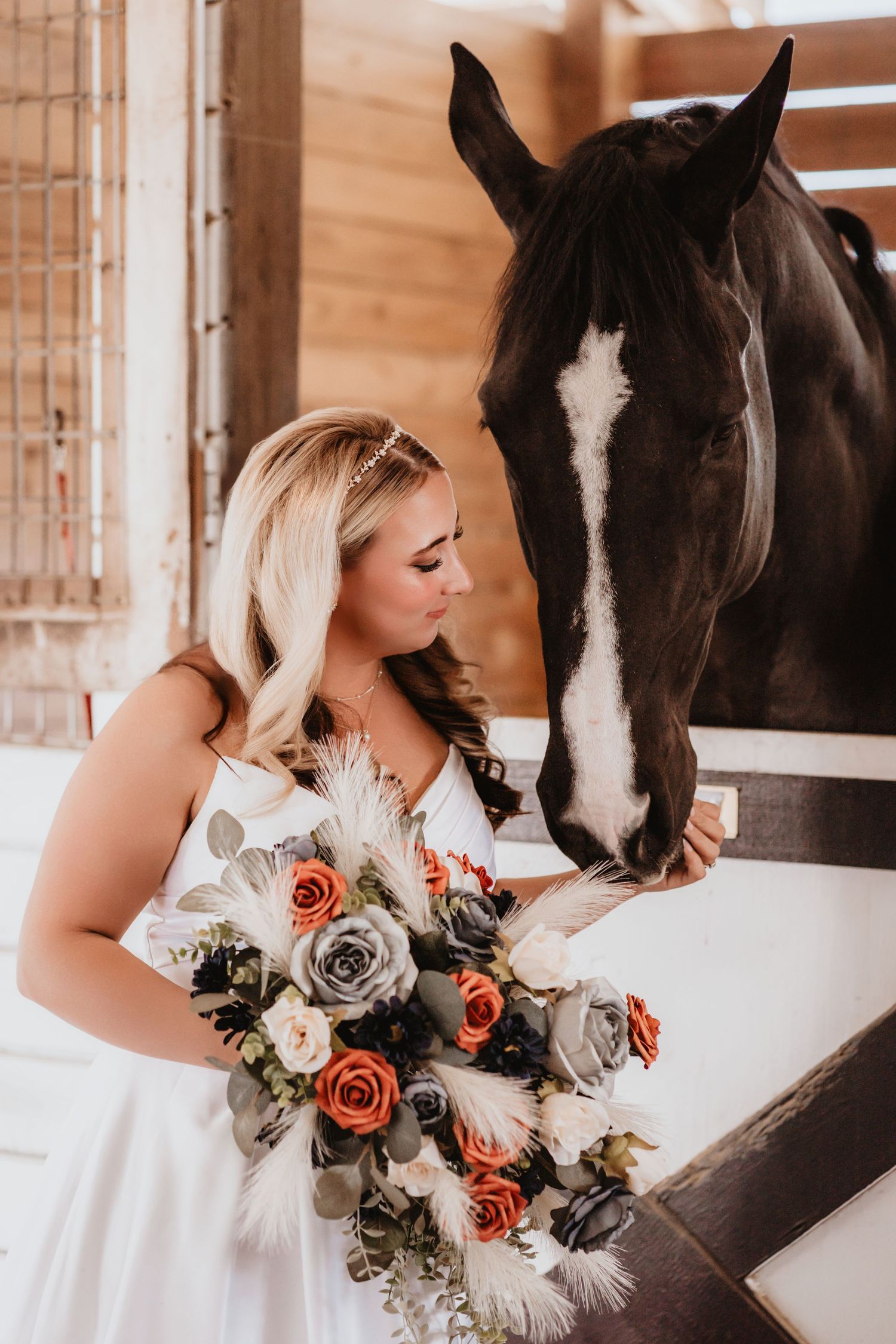 Bride in white dress gazes at a black horse while holding a terracotta rose bouquet.