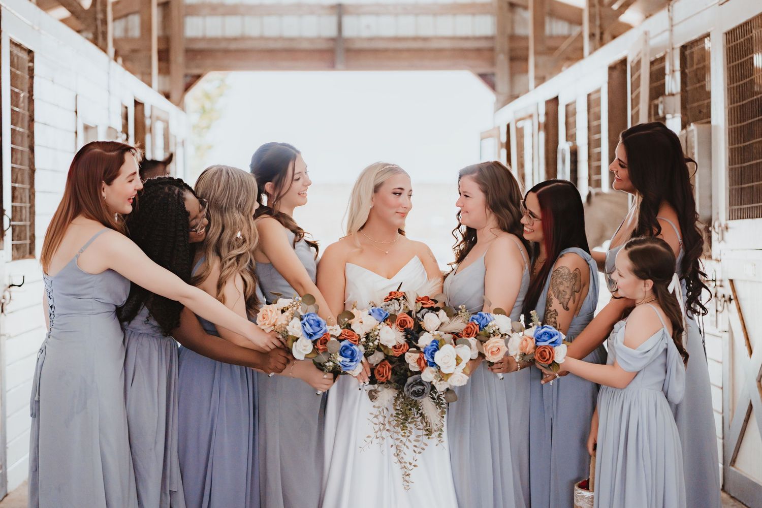 Bride surrounded by bridesmaids in blue-gray dresses laughing together in a rustic barn setting.