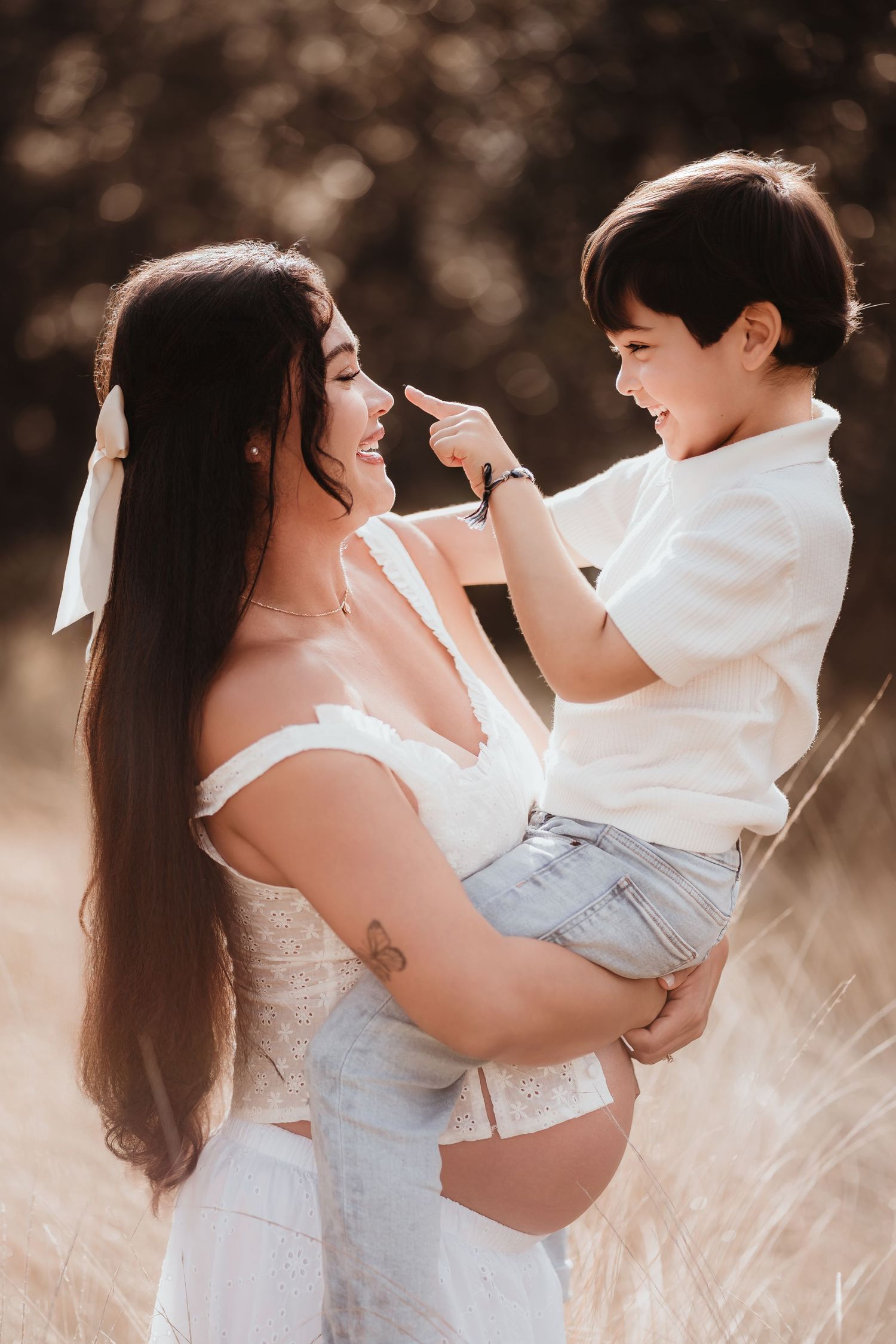 Pregnant woman with long dark hair laughing as young son playfully touches her nose outdoors.