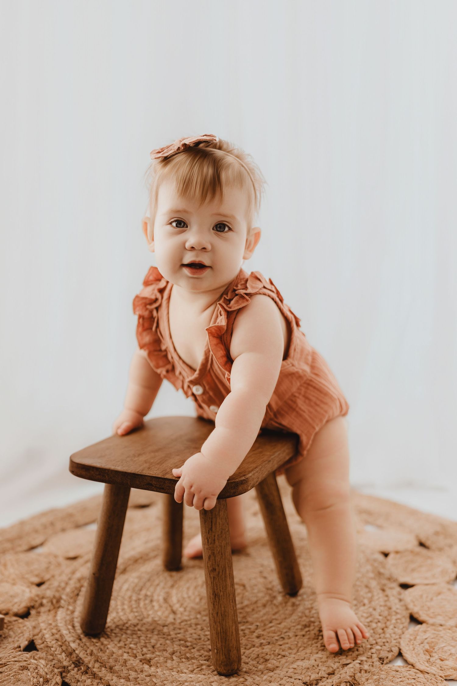 Baby girl in rust ruffle romper with bow headband leaning on small wooden stool on woven rug.