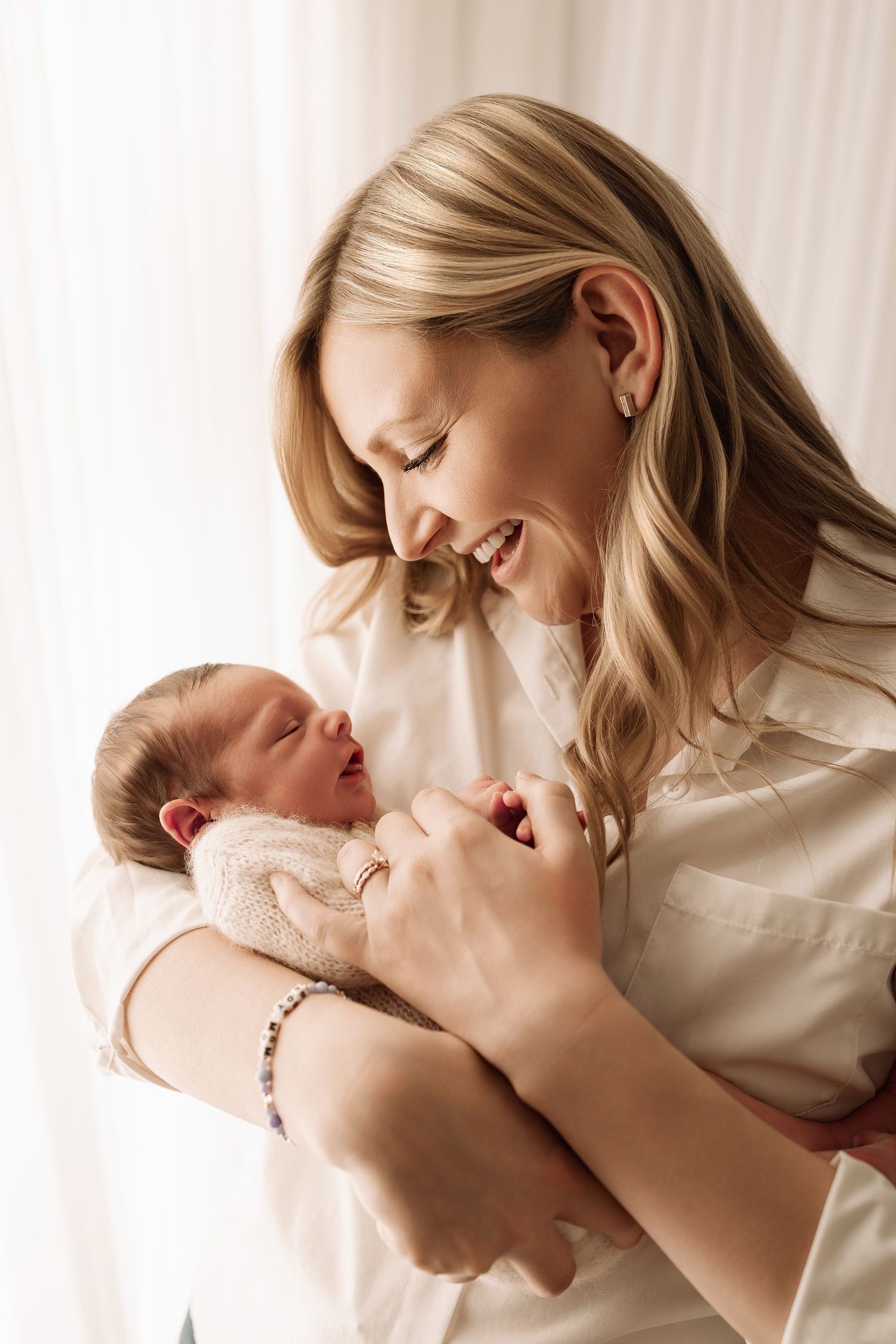 Smiling blonde mother laughing while holding her sleeping newborn baby in a bright white studio.