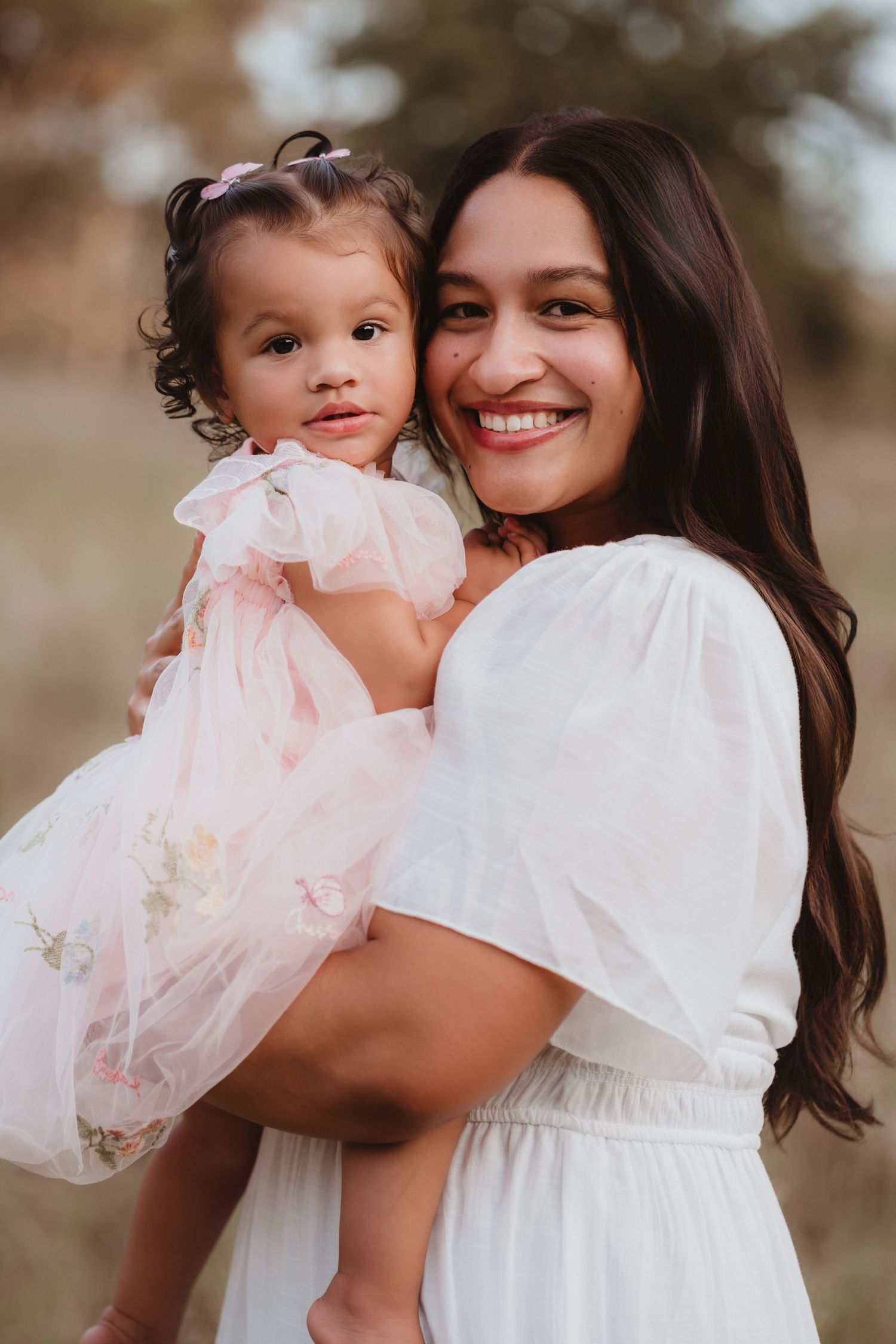 Smiling mother in white dress holding her young daughter in a pink floral tulle dress.