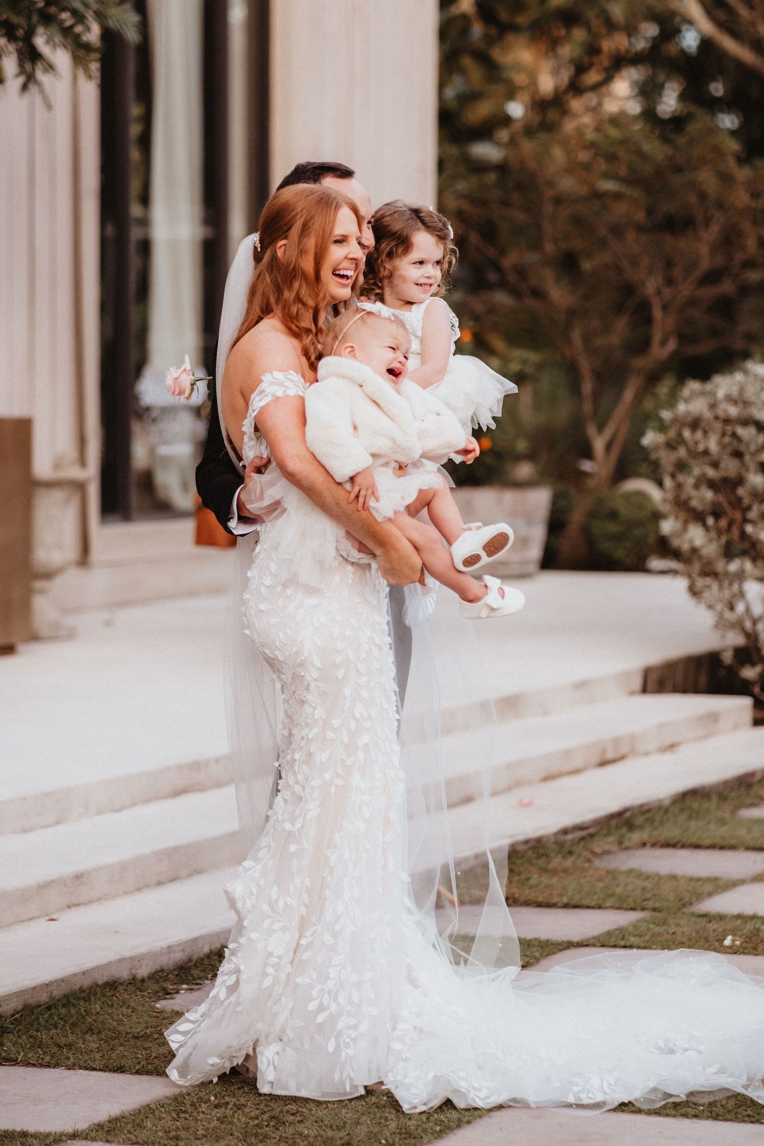 Laughing bride in fitted lace wedding gown holding two young children in white outfits outdoors.