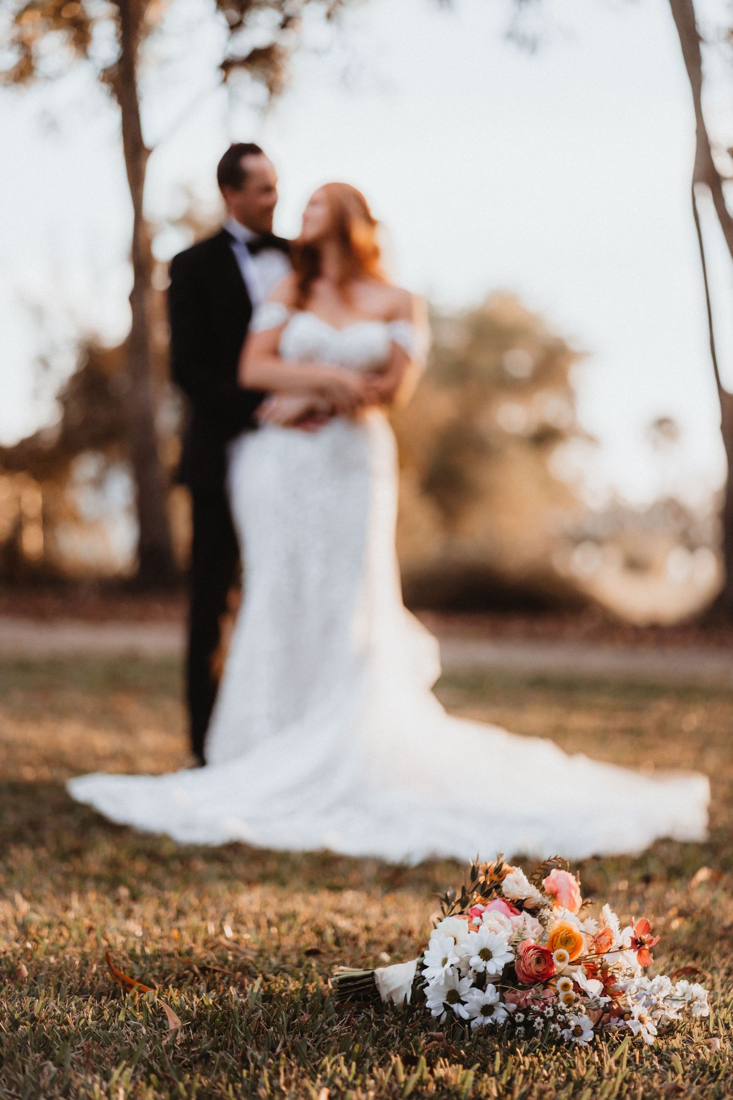 Colorful bridal bouquet of daisies and roses in foreground with bride and groom embracing at sunset.